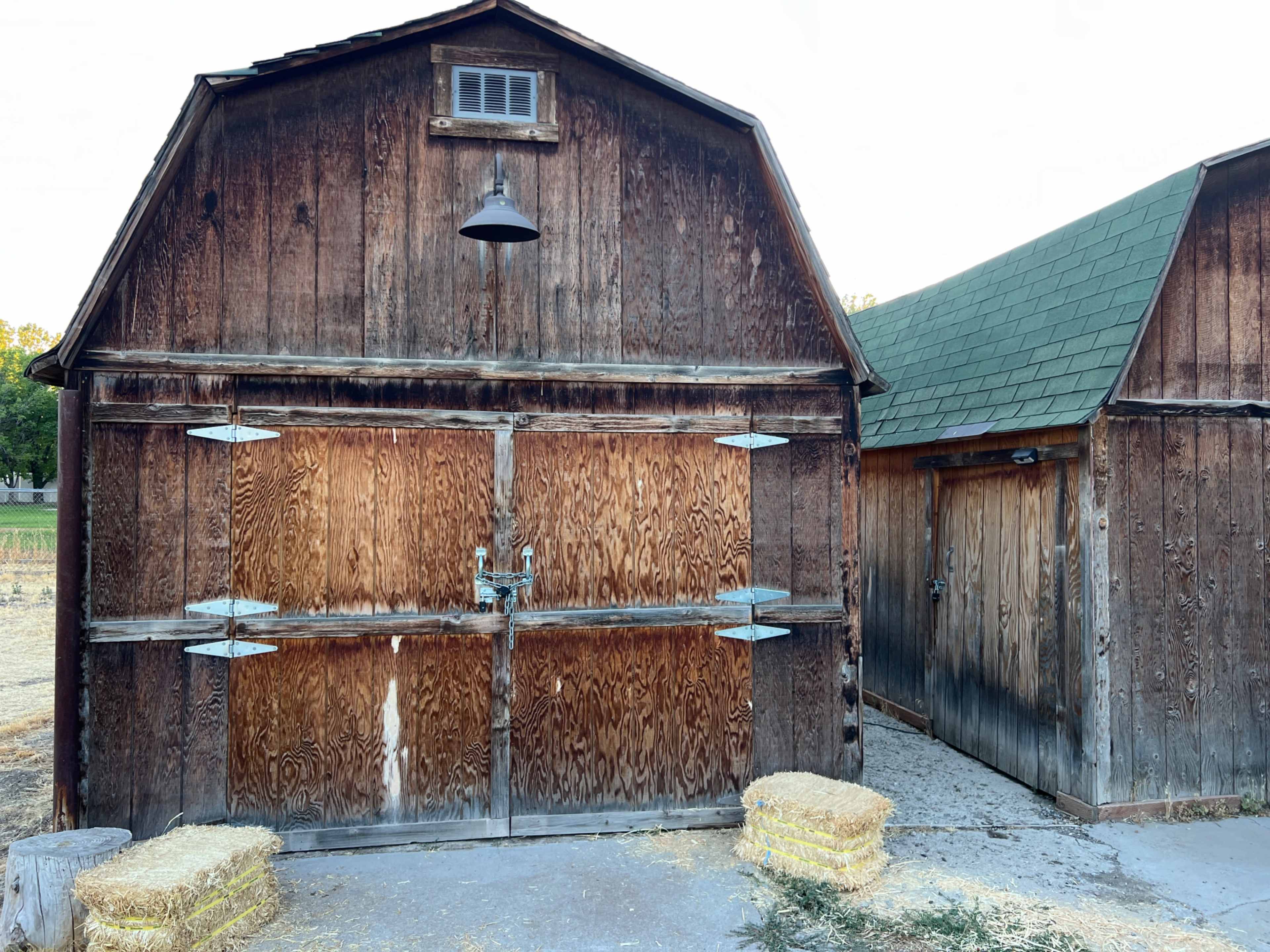 The image shows a weathered wooden barn with double doors and a smaller attached structure, both featuring rustic designs and surrounded by bales of hay on the ground.
