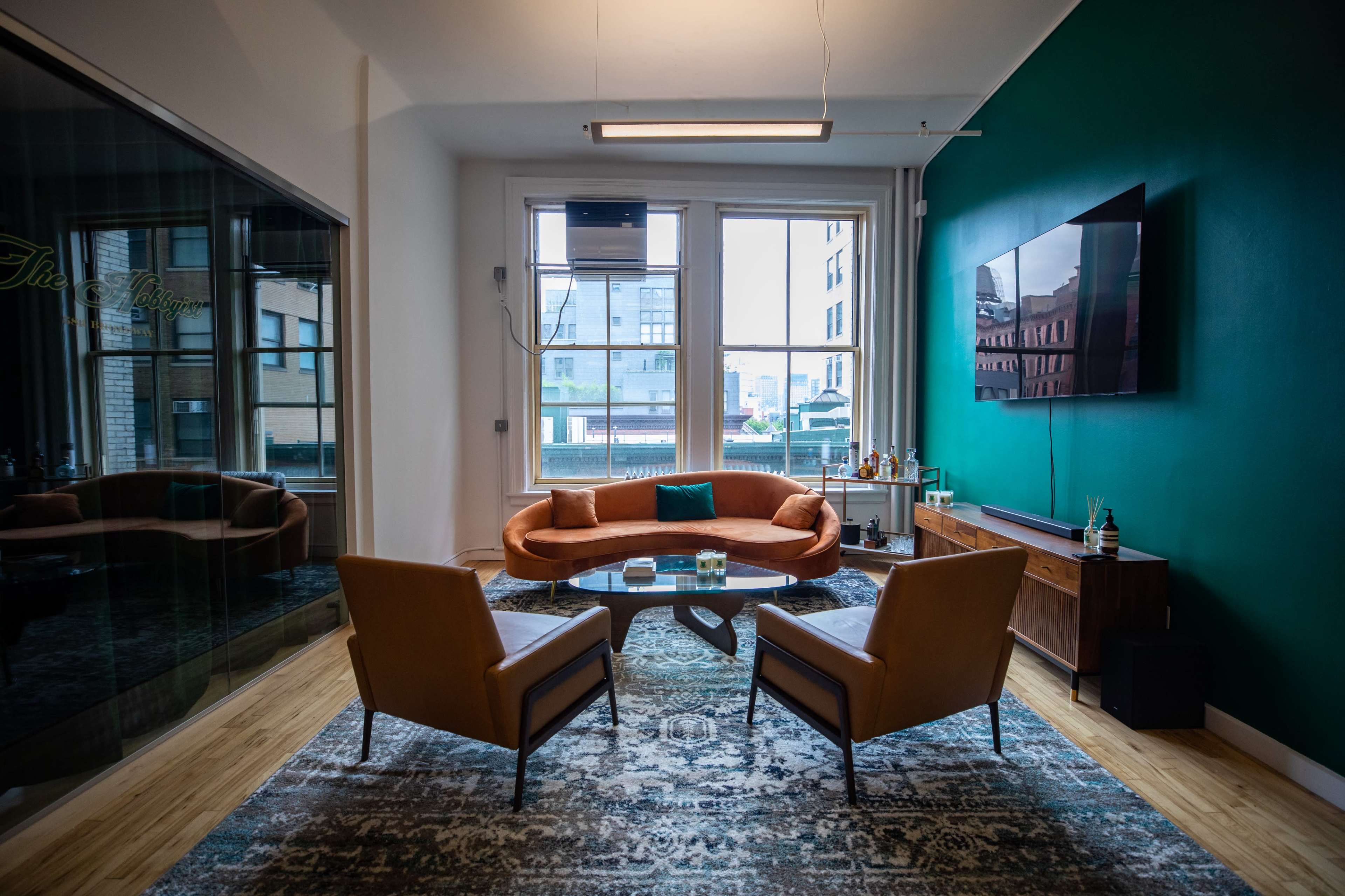 A modern living room with a curved orange sofa, two brown chairs, a coffee table, and a wall-mounted television, all set against a green accent wall and large windows.