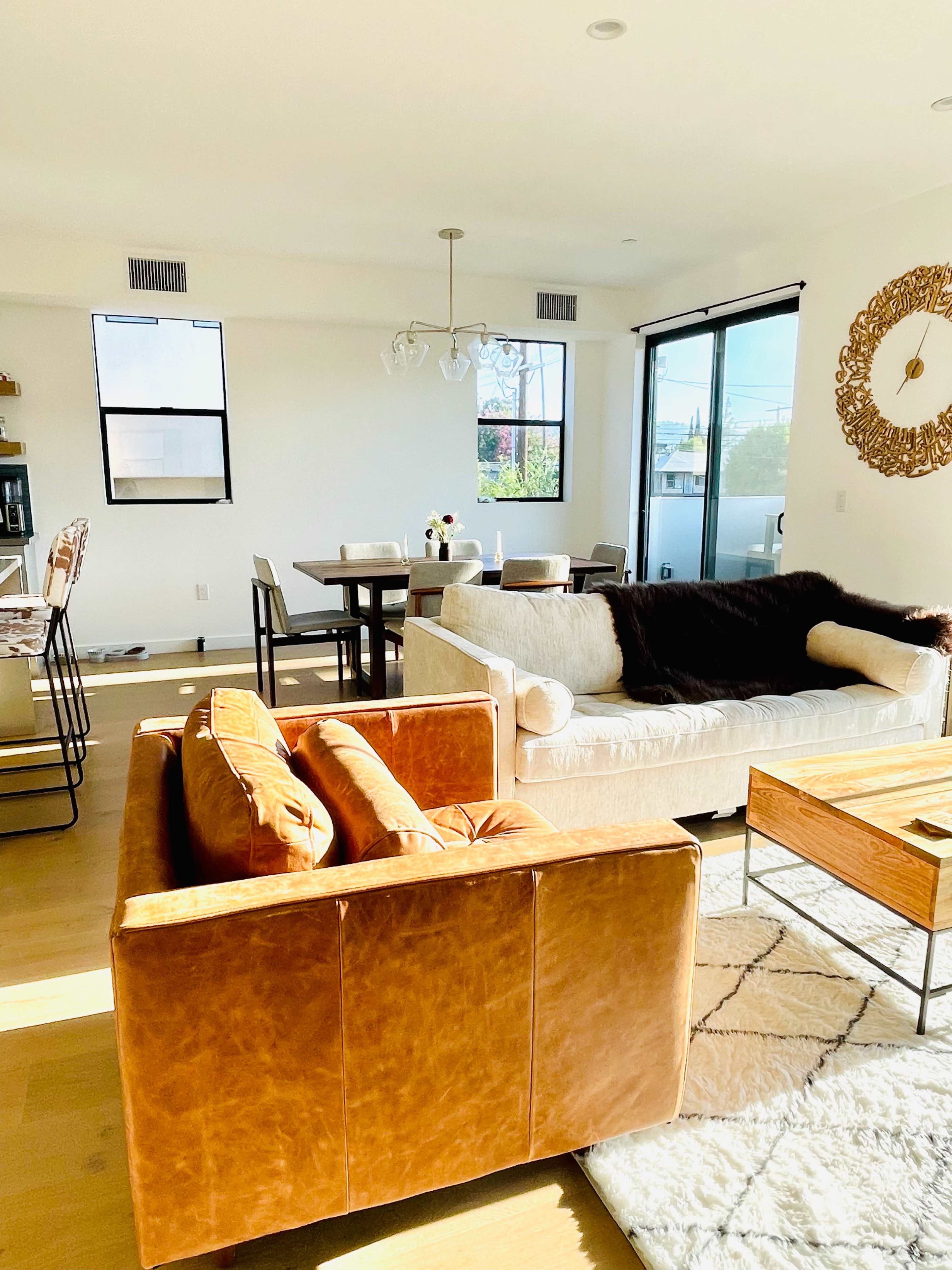 A modern living room featuring a leather sofa, a light-colored couch, a wooden coffee table, and a dining area with a table and chairs under natural light from large windows.