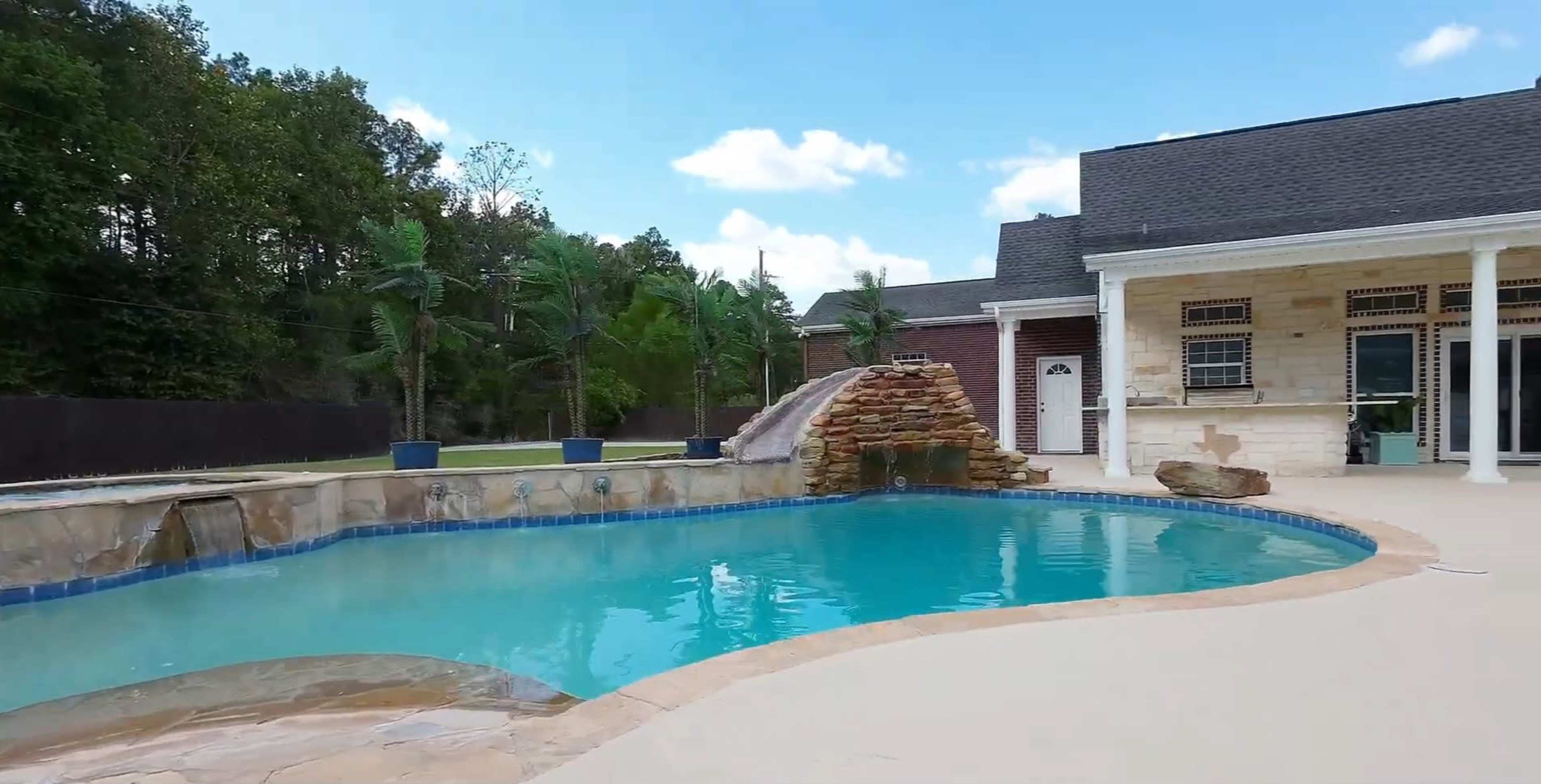 The image depicts a swimming pool with a stone waterfall, surrounded by palm trees and a spacious patio next to a house.