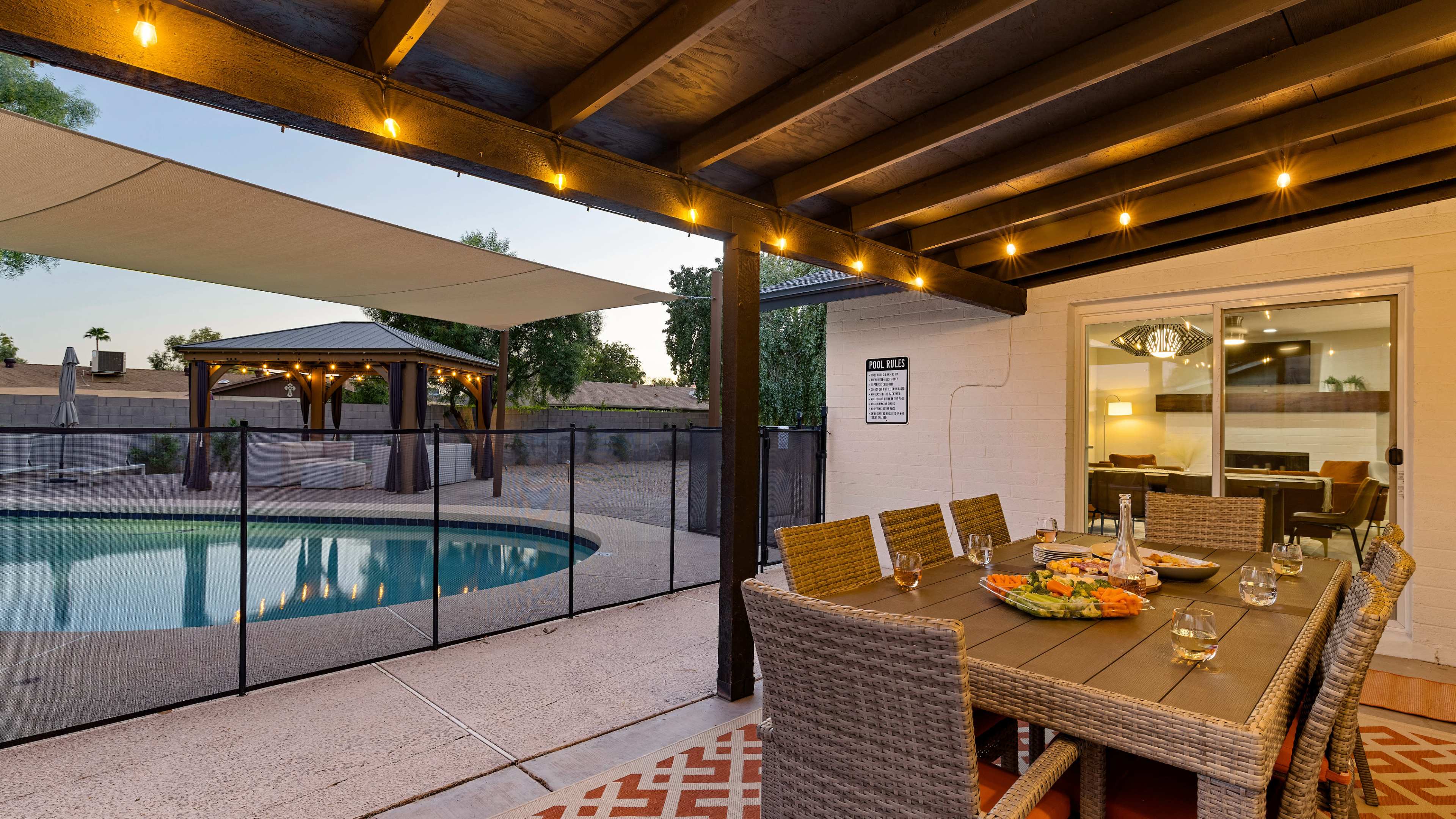 A dining table with chairs is set up under a covered patio beside a swimming pool, with a small gazebo visible in the background.
