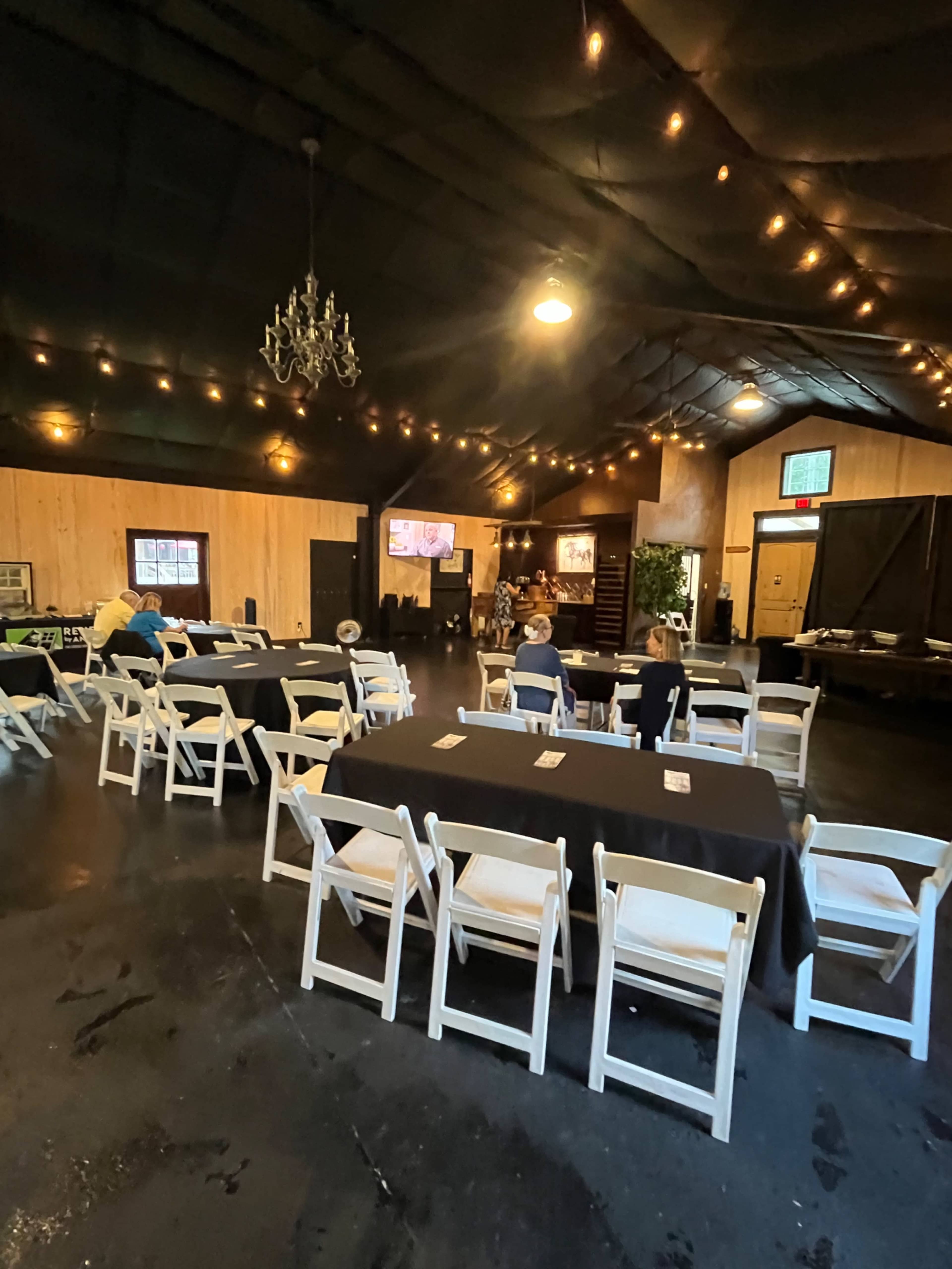 The image shows a spacious indoor venue with rows of white folding chairs and black tables, illuminated by soft overhead lights and a chandelier.
