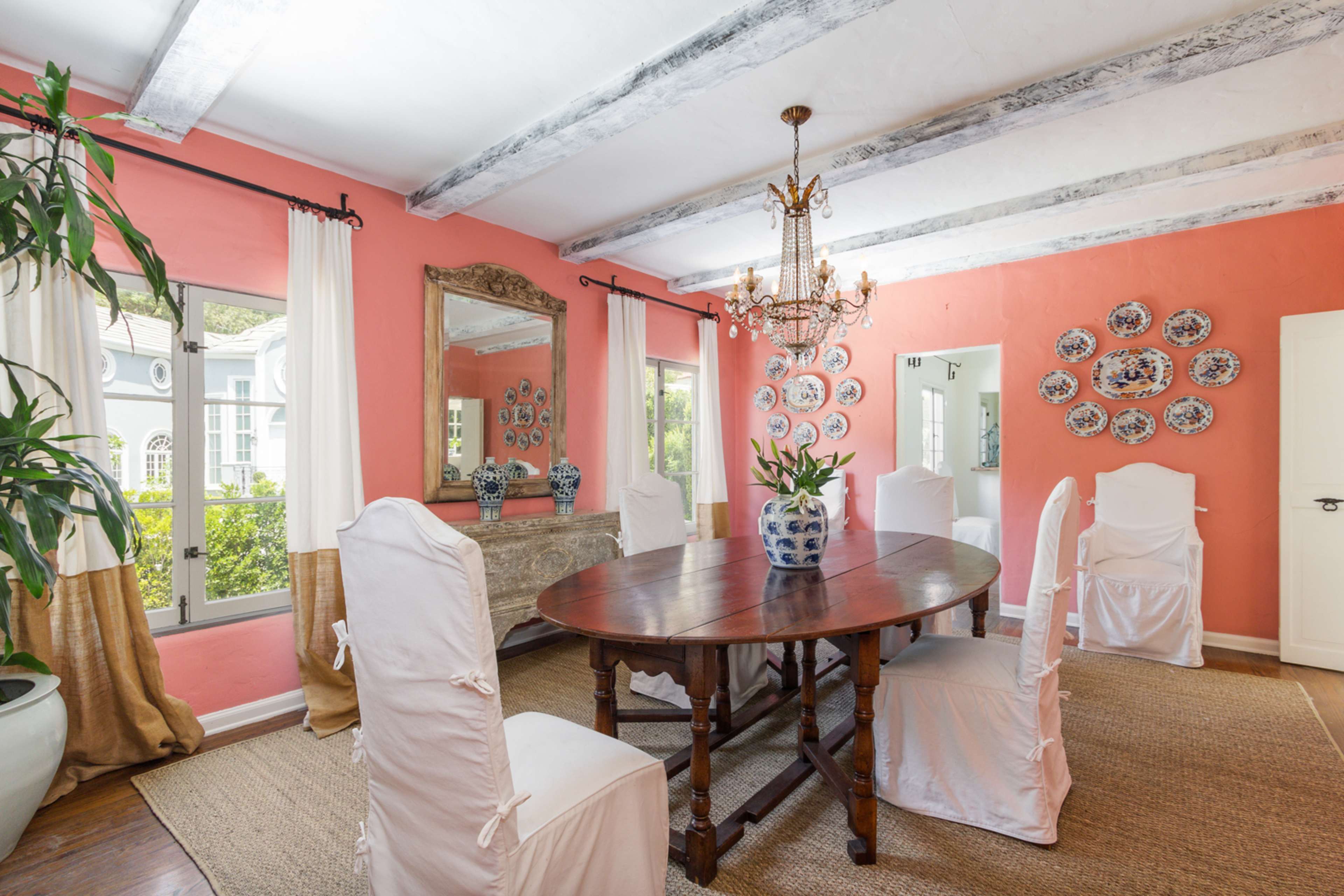 The dining room features a round wooden table surrounded by six white slipcovered chairs, with pink walls, a chandelier, and decorative plates on display.
