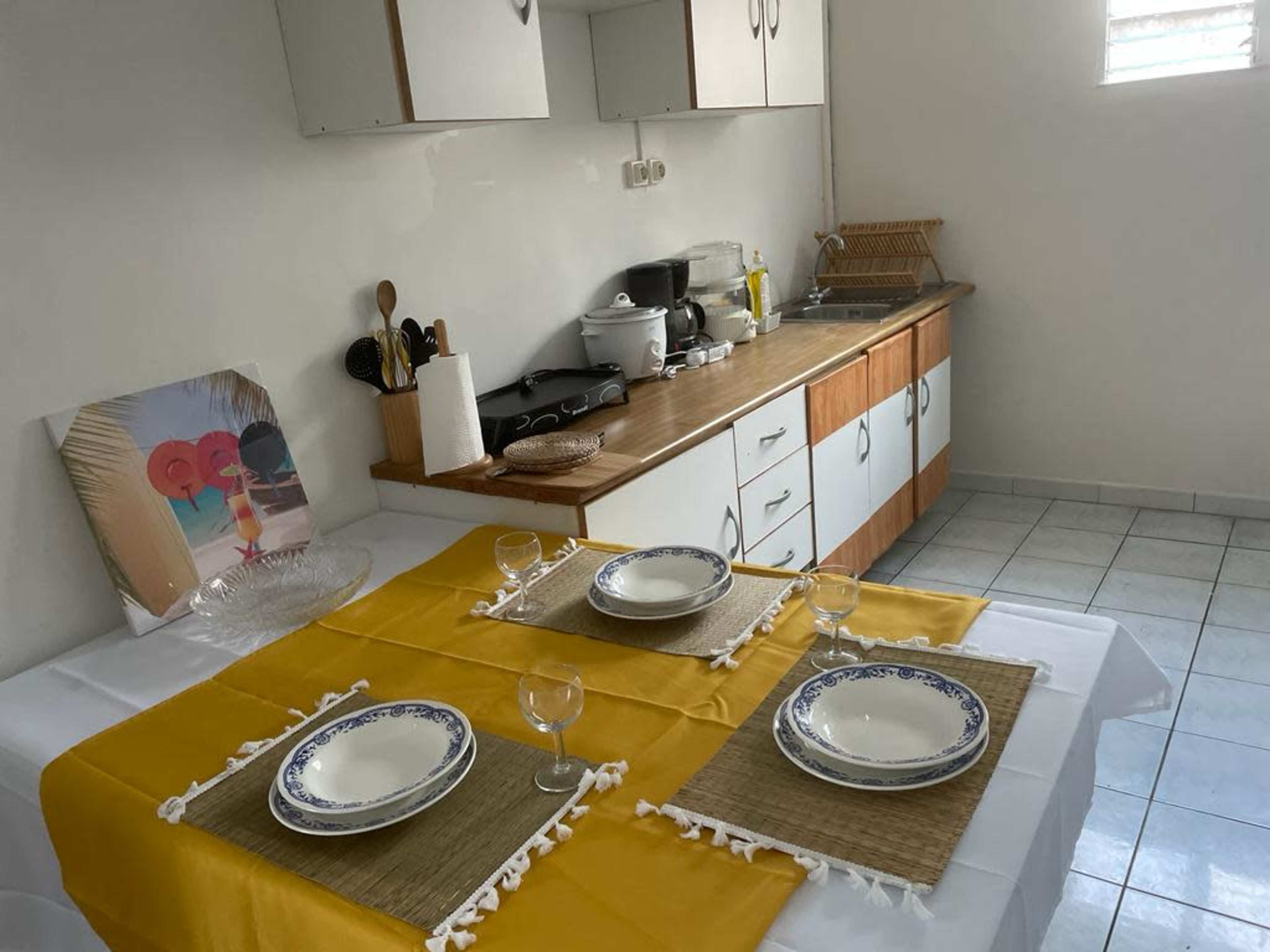 A kitchen is set with a yellow tablecloth, four plates, and glassware ready for a meal, with utensils and appliances visible in the background.