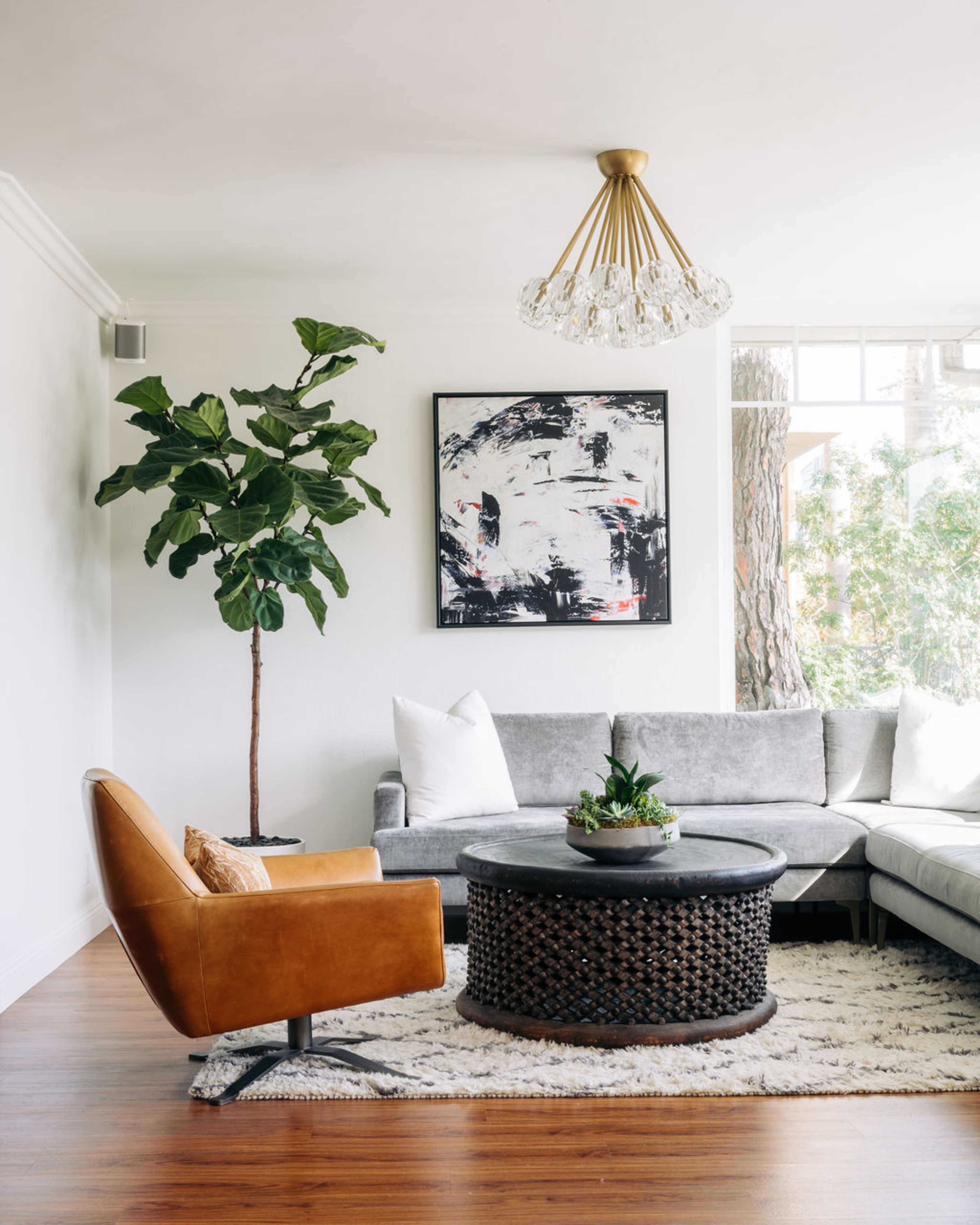 A modern living room features a gray sectional sofa, a brown accent chair, a round coffee table with a woven design, and a tall potted plant, all under a chandelier.