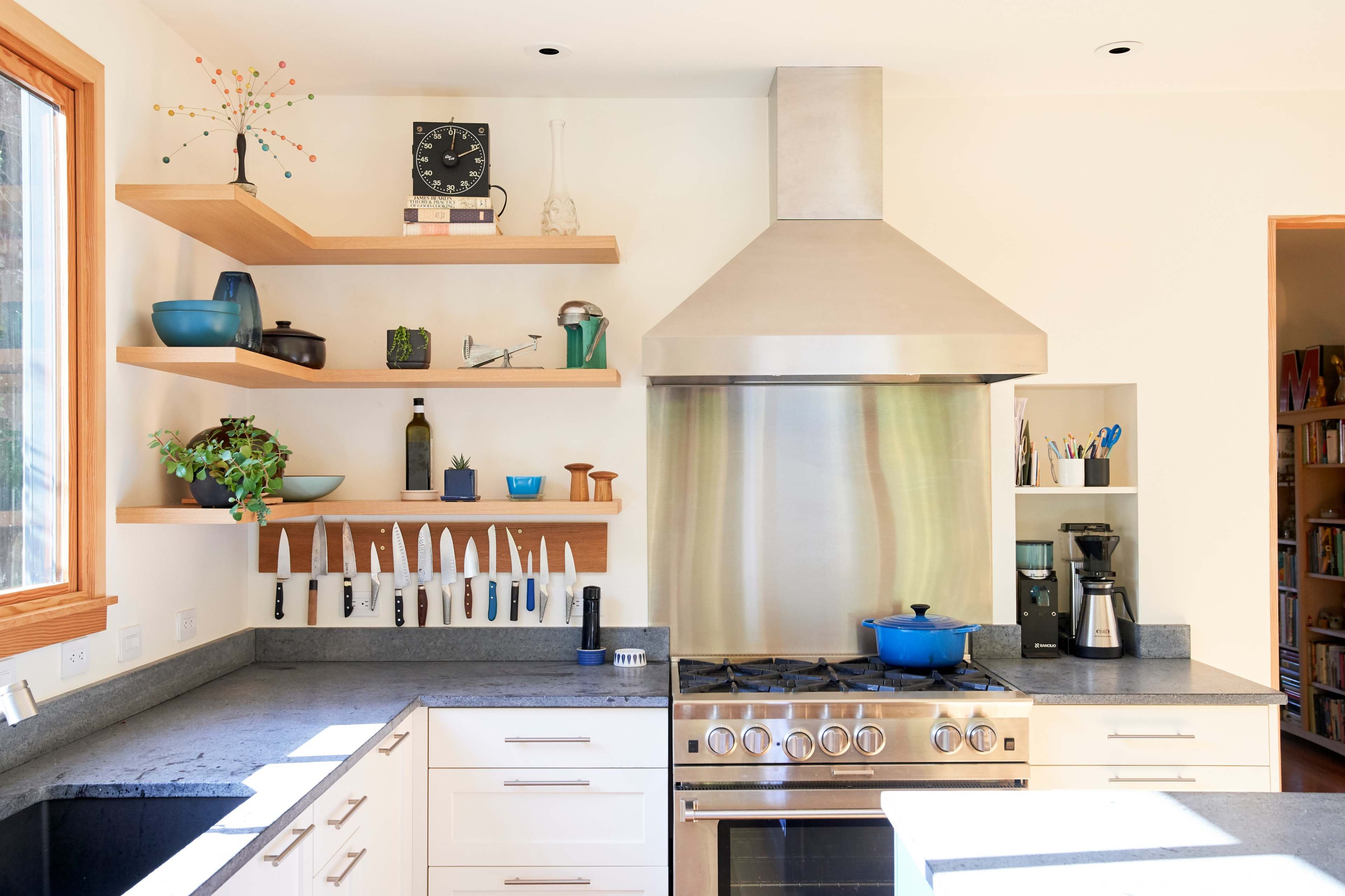 A modern kitchen features stainless steel appliances, a large ventilation hood, open wooden shelves, and a variety of cooking utensils neatly arranged.