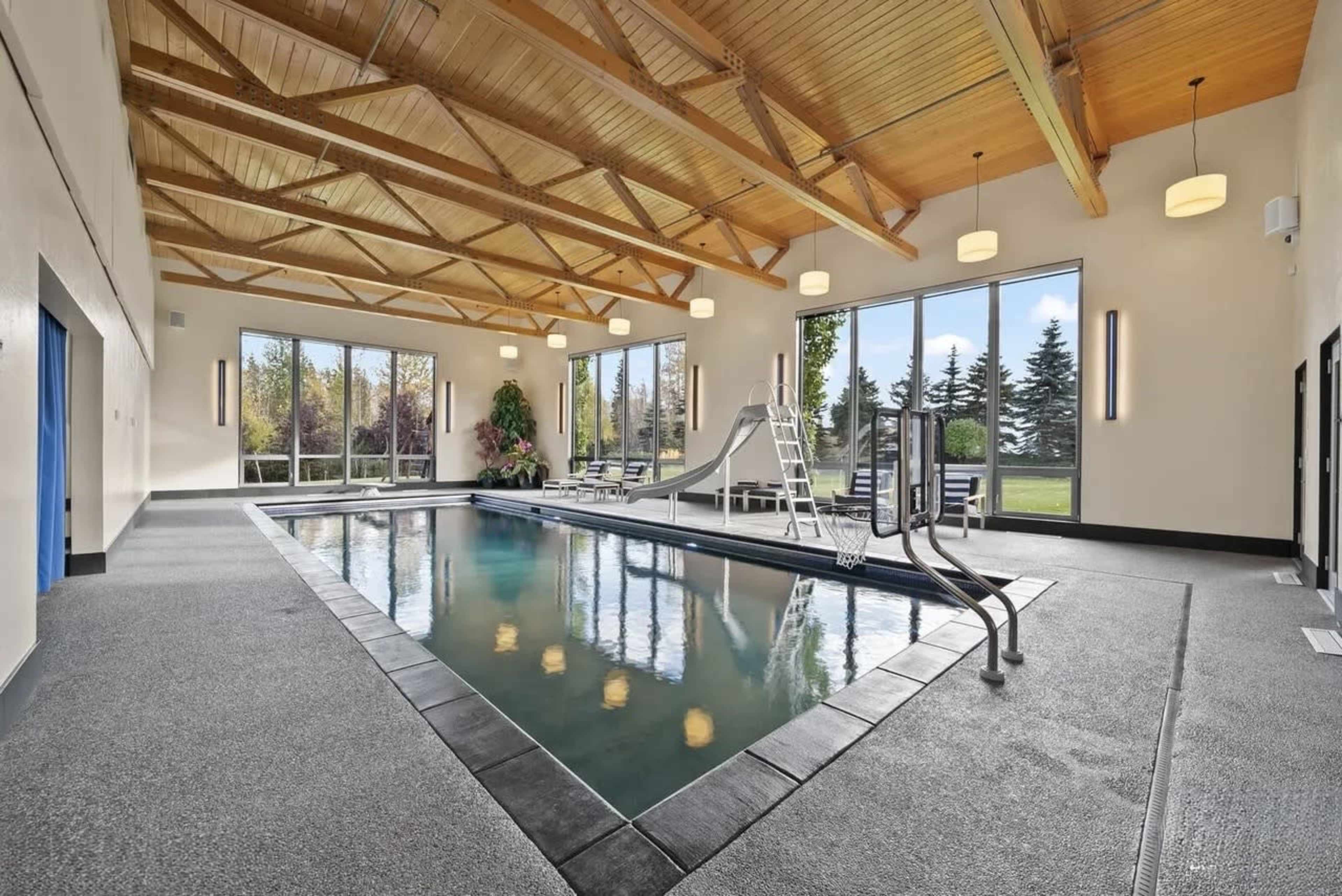 The image shows an indoor swimming pool area with large windows, wooden beams overhead, and a slide next to the pool.