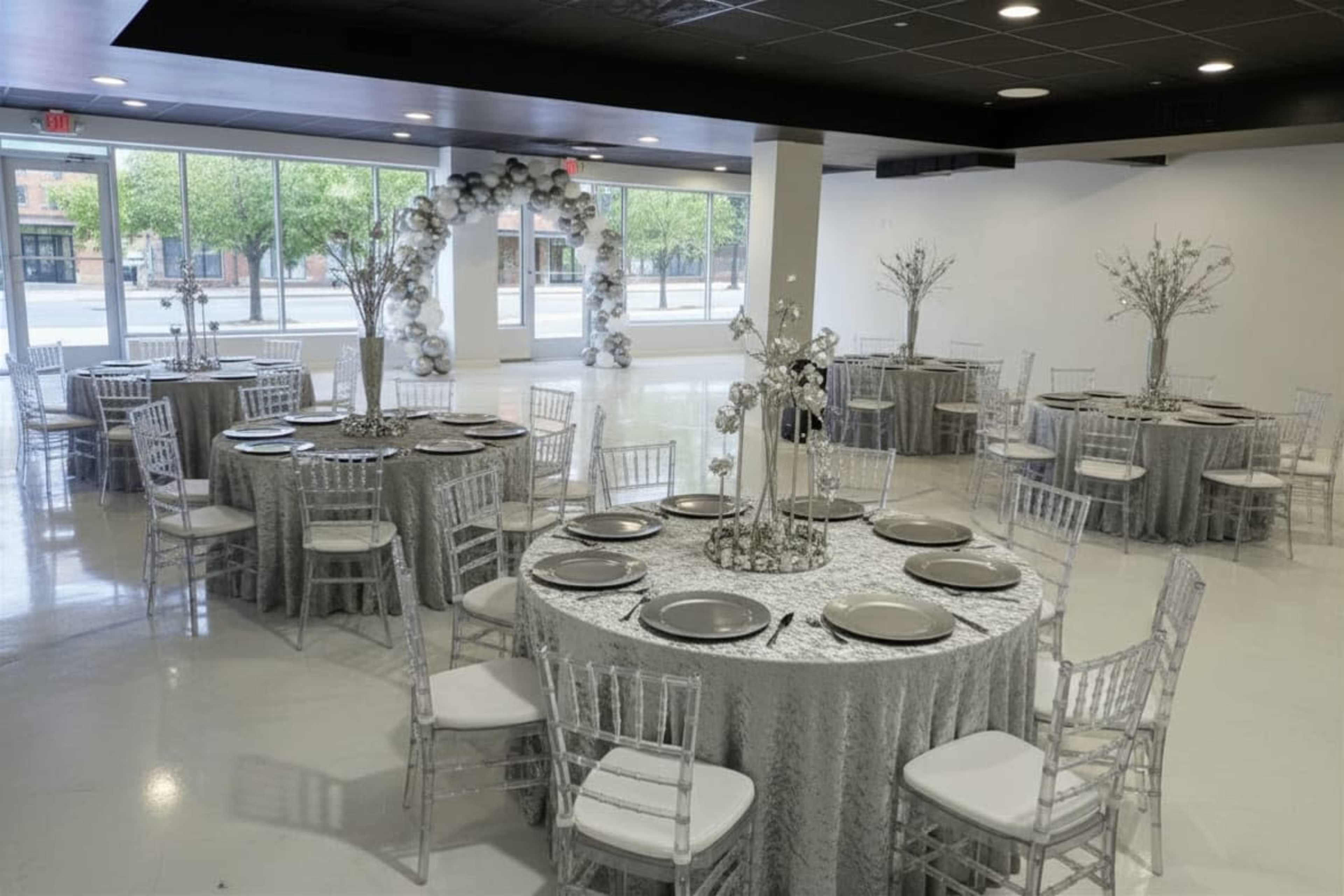 A spacious event hall features elegantly set tables with silver tablecloths, each adorned with white dinnerware and surrounded by transparent chairs.