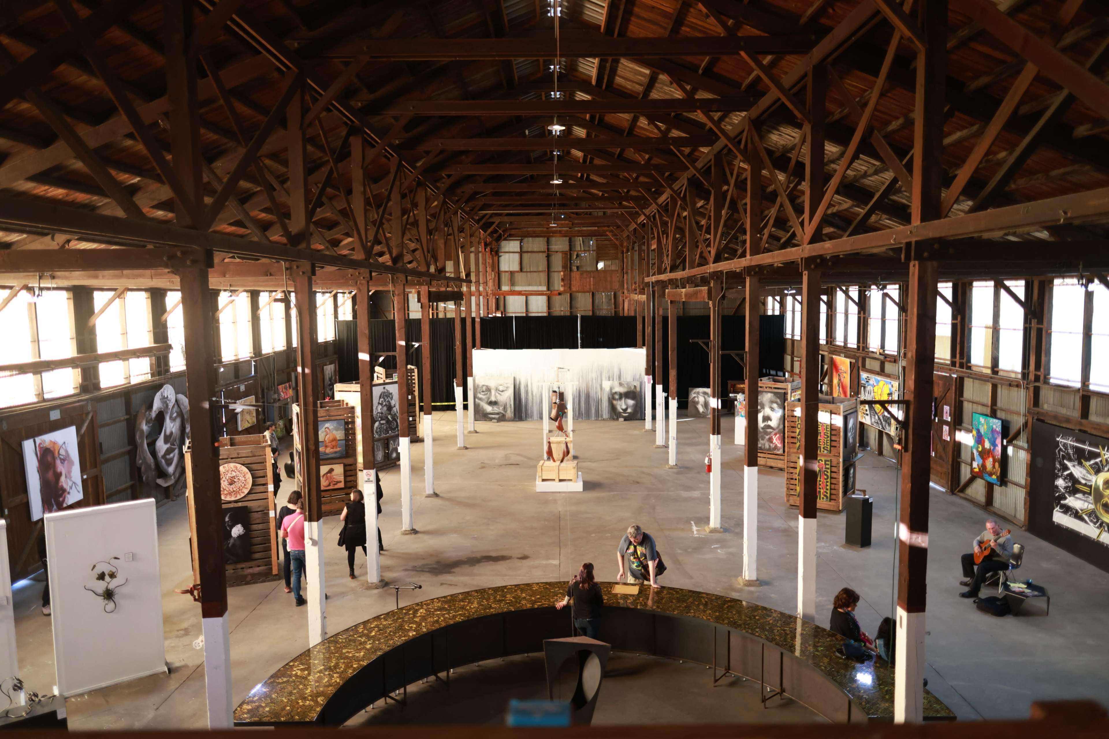The image shows an expansive art gallery housed in a rustic wooden building, featuring various artworks displayed on walls and pedestals.