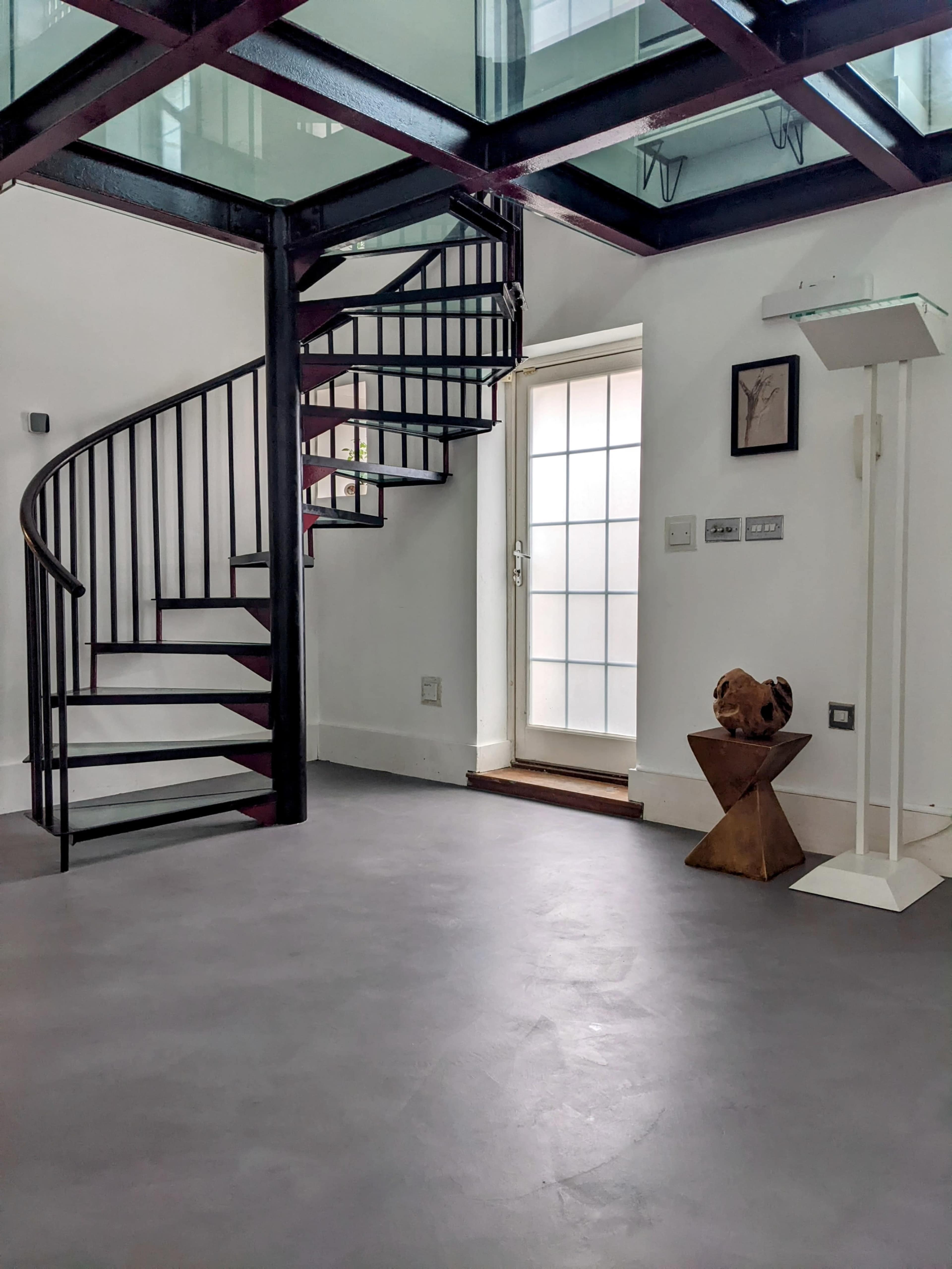 A modern interior featuring a spiral staircase on the left, a glass ceiling above, and a geometric sculpture near a doorway.