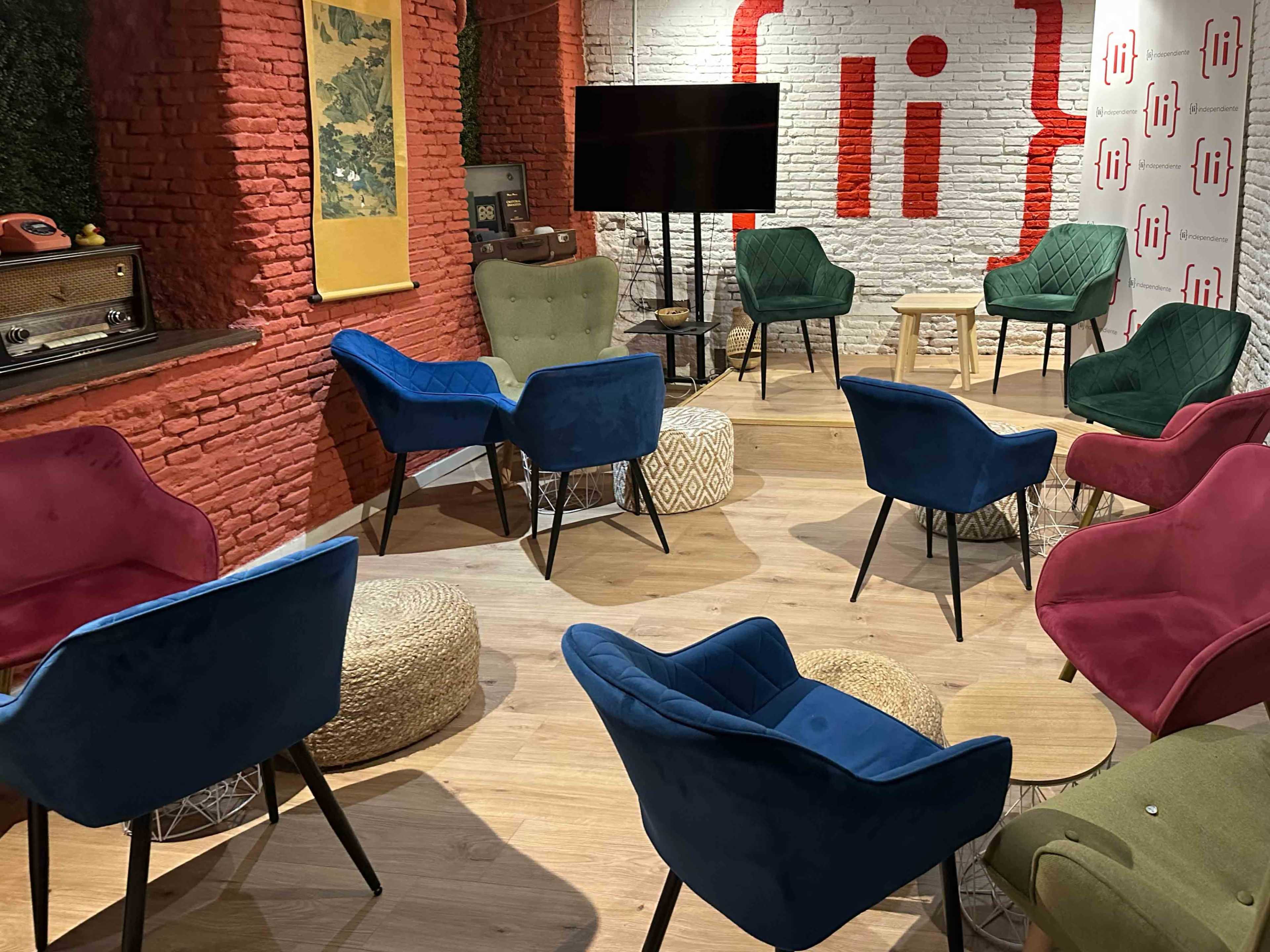 The image shows a cozy meeting space with colorful upholstered chairs arranged around a small wooden table, set against a backdrop of red brick walls and a television.