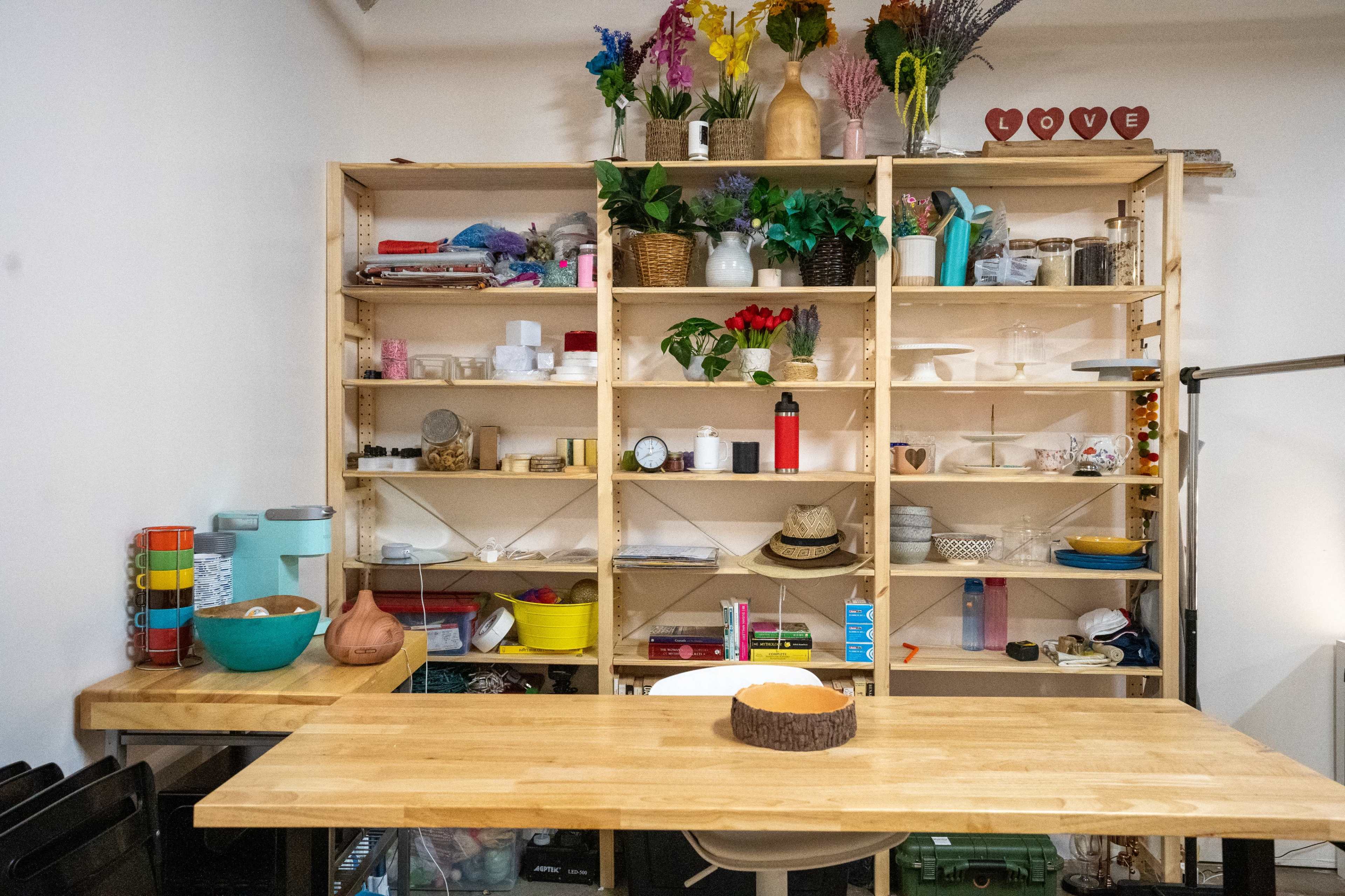 A wooden table in front of a bookshelf filled with various items, including plants, containers, and decorative objects, in a room with neutral walls.