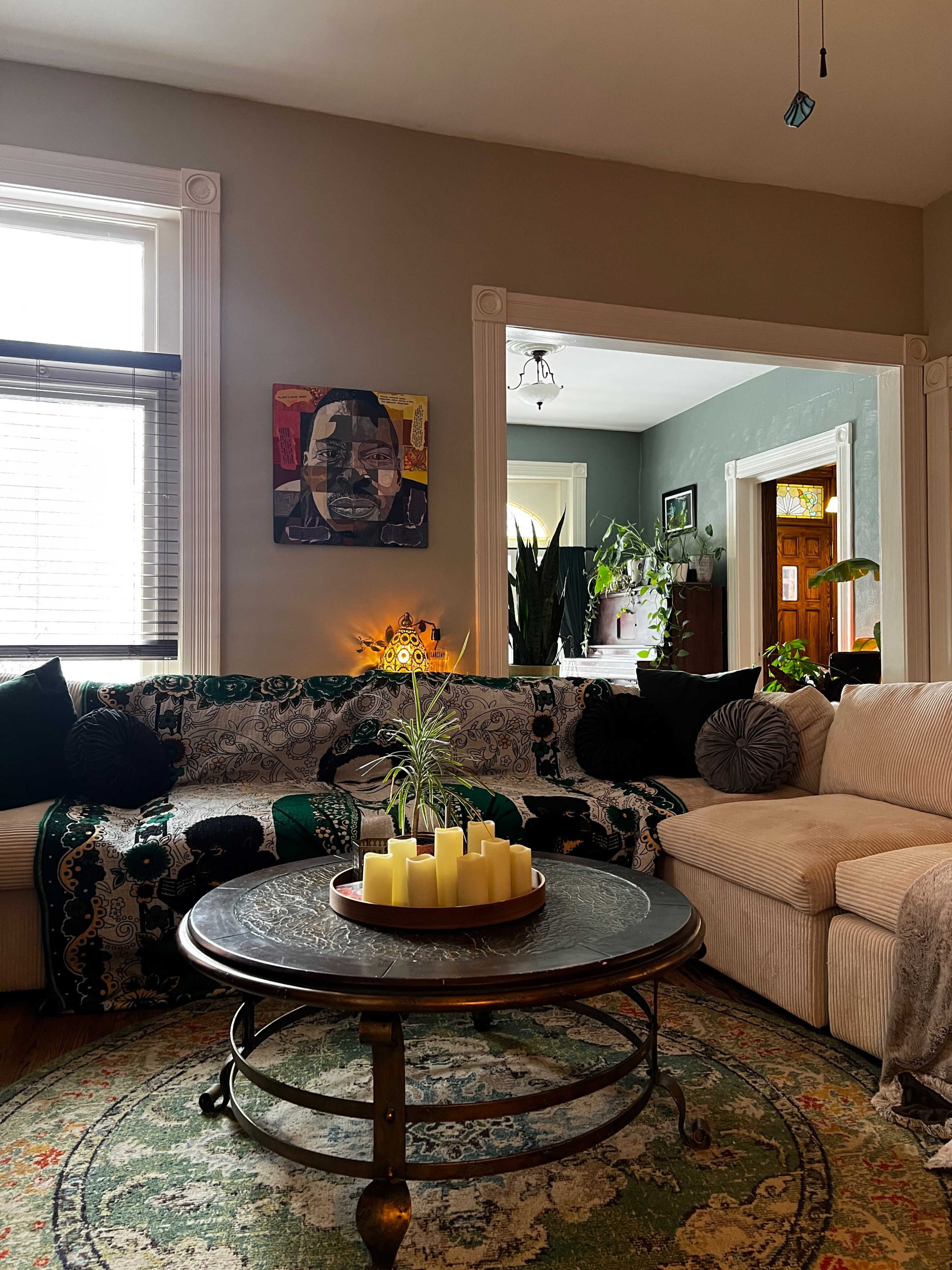 The living room features a patterned couch adorned with dark pillows, a round coffee table with yellow candles, and a colorful rug under a wall displaying art and houseplants.
