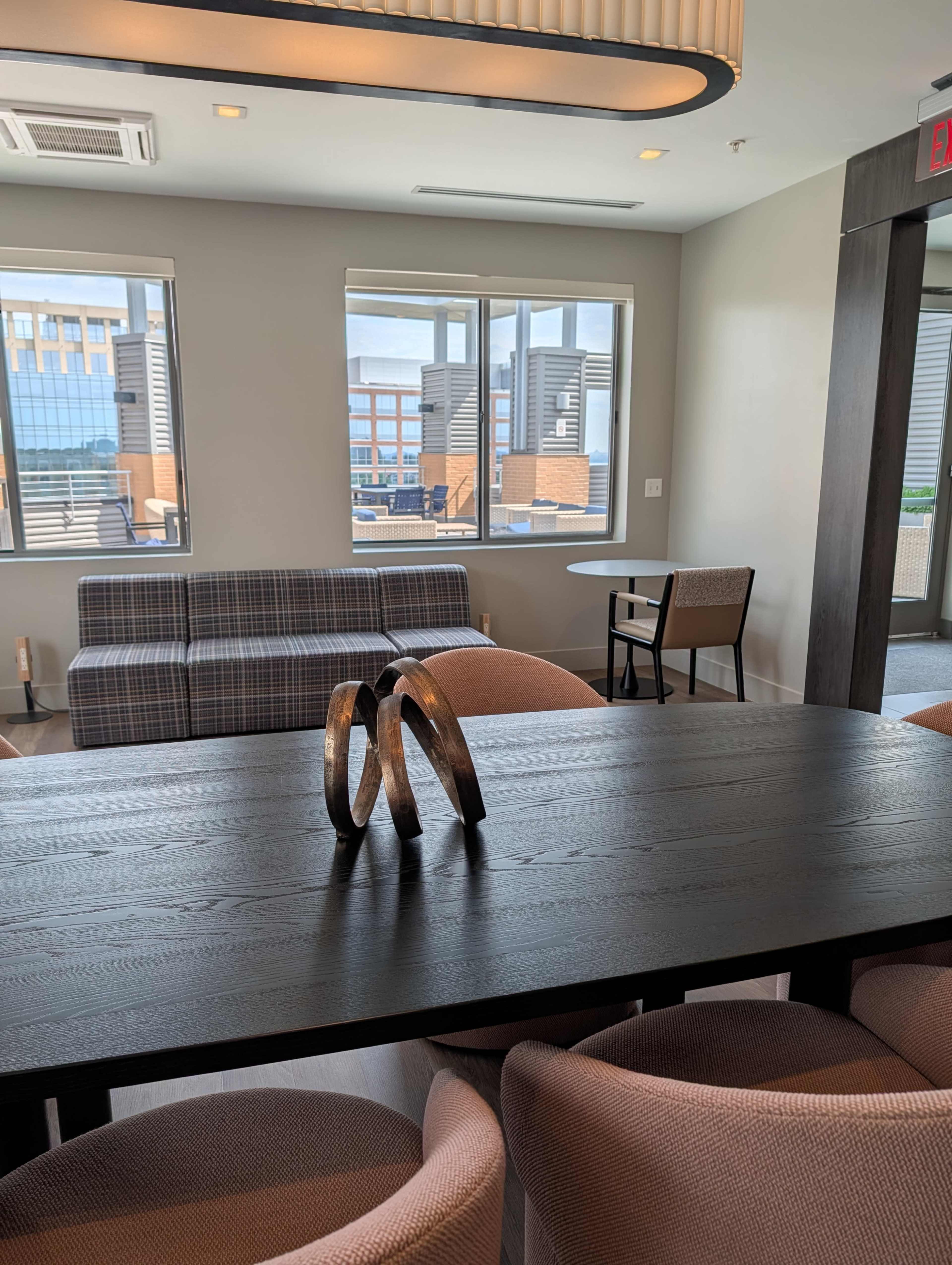 The image shows a modern interior space featuring a dining table with decorative rings, plush chairs, a sectional sofa, and large windows revealing a view outside.