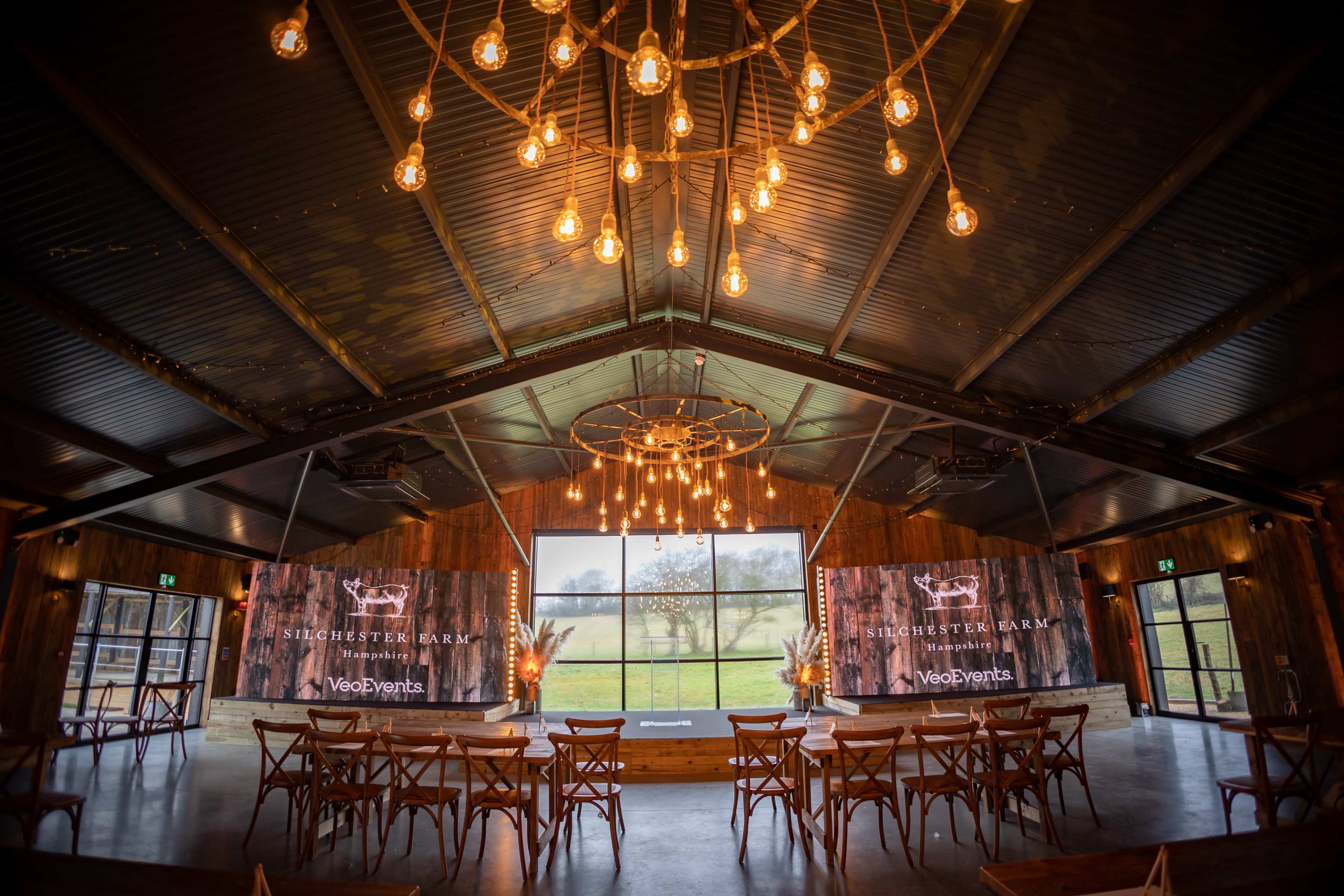 The interior of a venue features wooden walls, long tables, and illuminated chandeliers, with large windows overlooking a landscape.