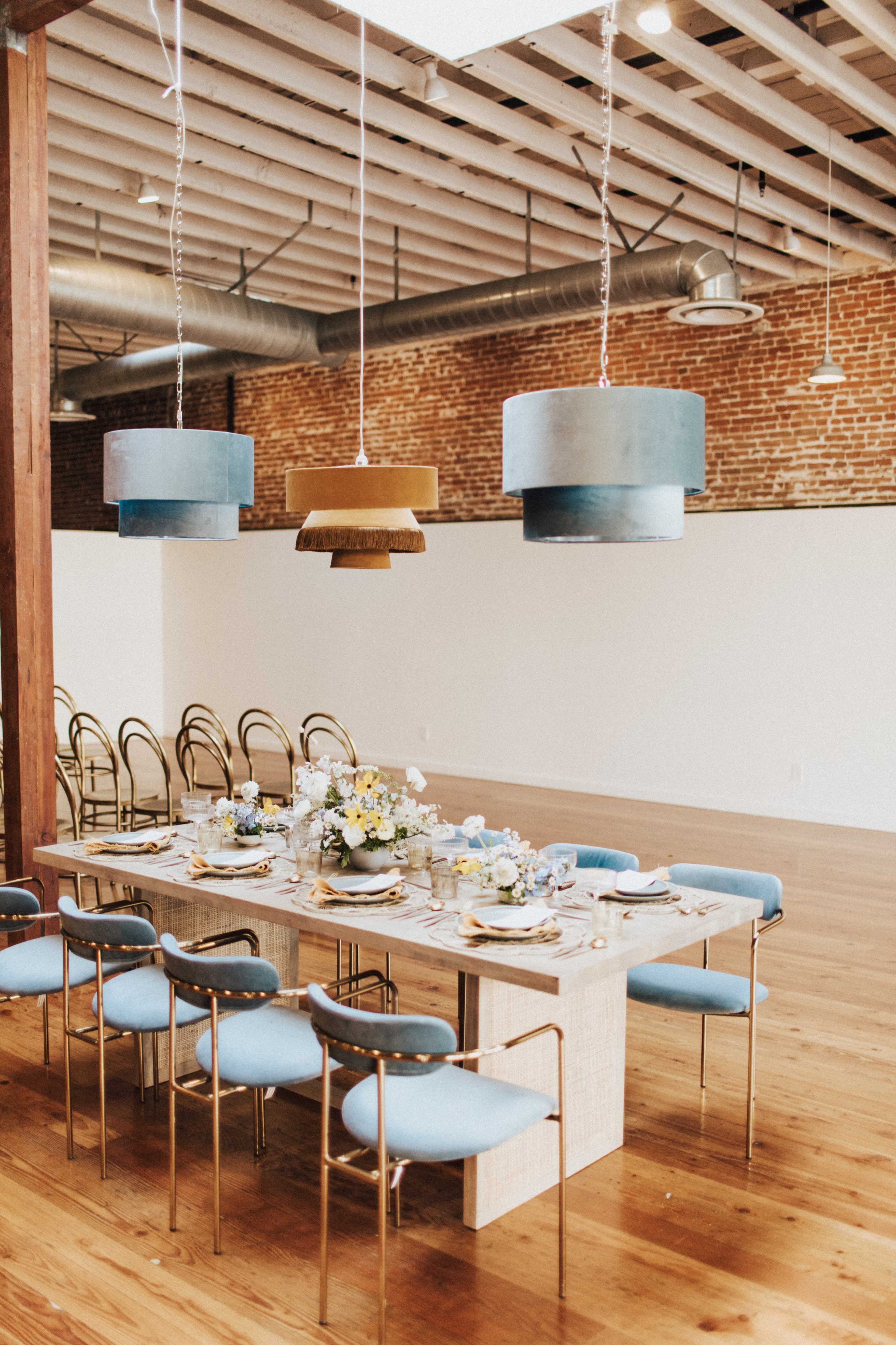 A long dining table with blue chairs is set with plates and flowers, beneath three decorative light fixtures in a spacious room with exposed brick walls and wooden floors.