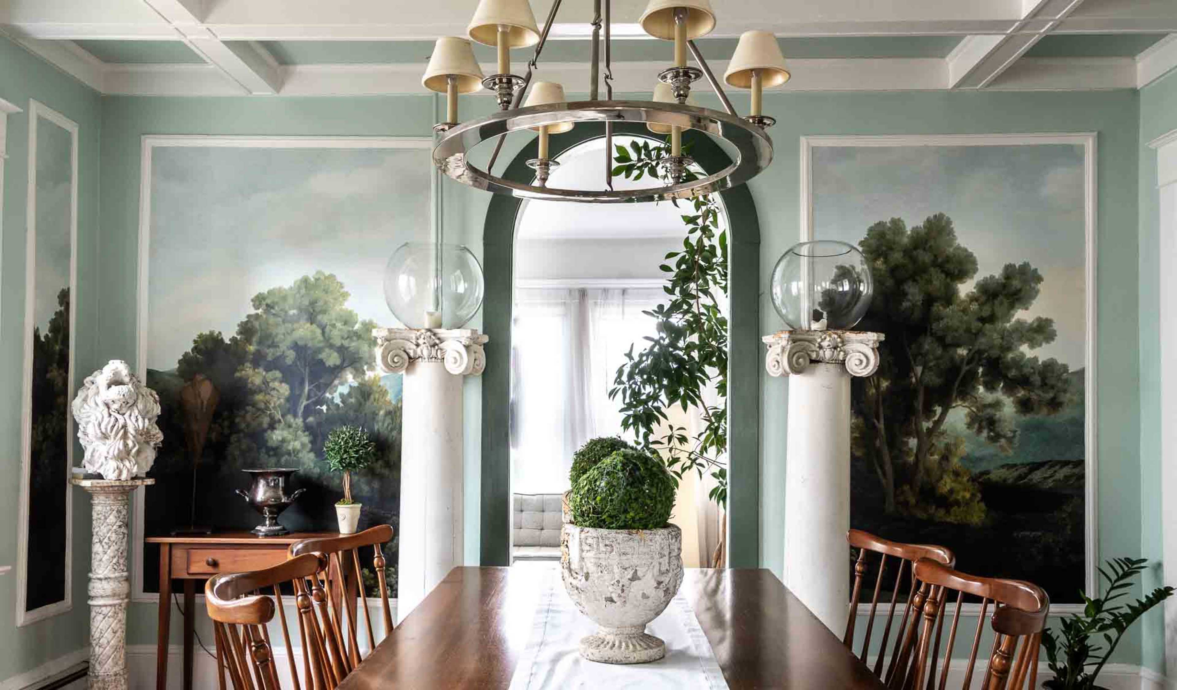 The image shows a dining room with a long wooden table, surrounded by chairs, and adorned with wall murals of trees and greenery, a chandelier overhead, and decorative plants in the corners.