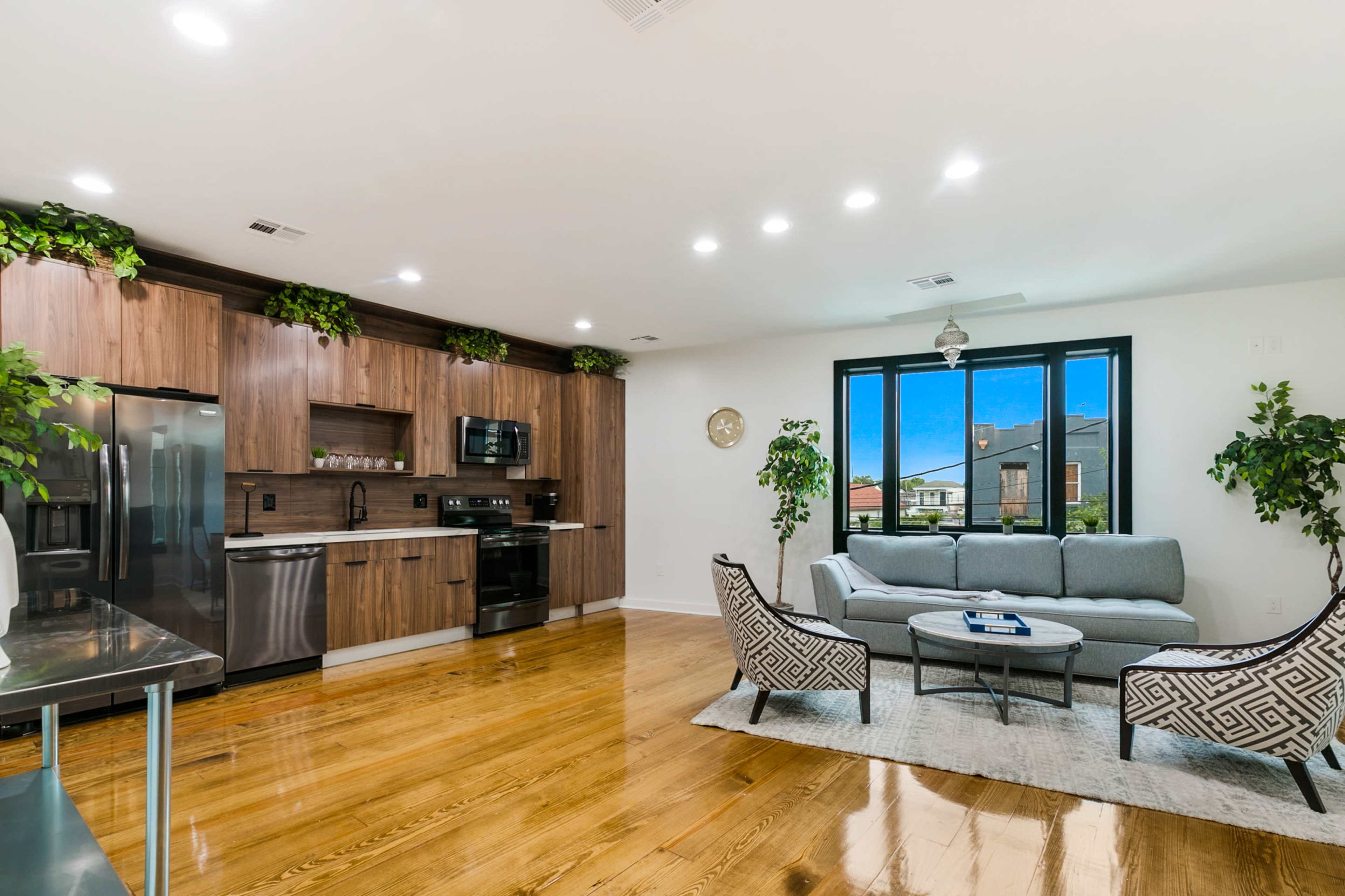 The image shows a modern living space with a kitchen and a seating area featuring a gray sofa and patterned armchairs against wooden cabinetry and polished hardwood floors.