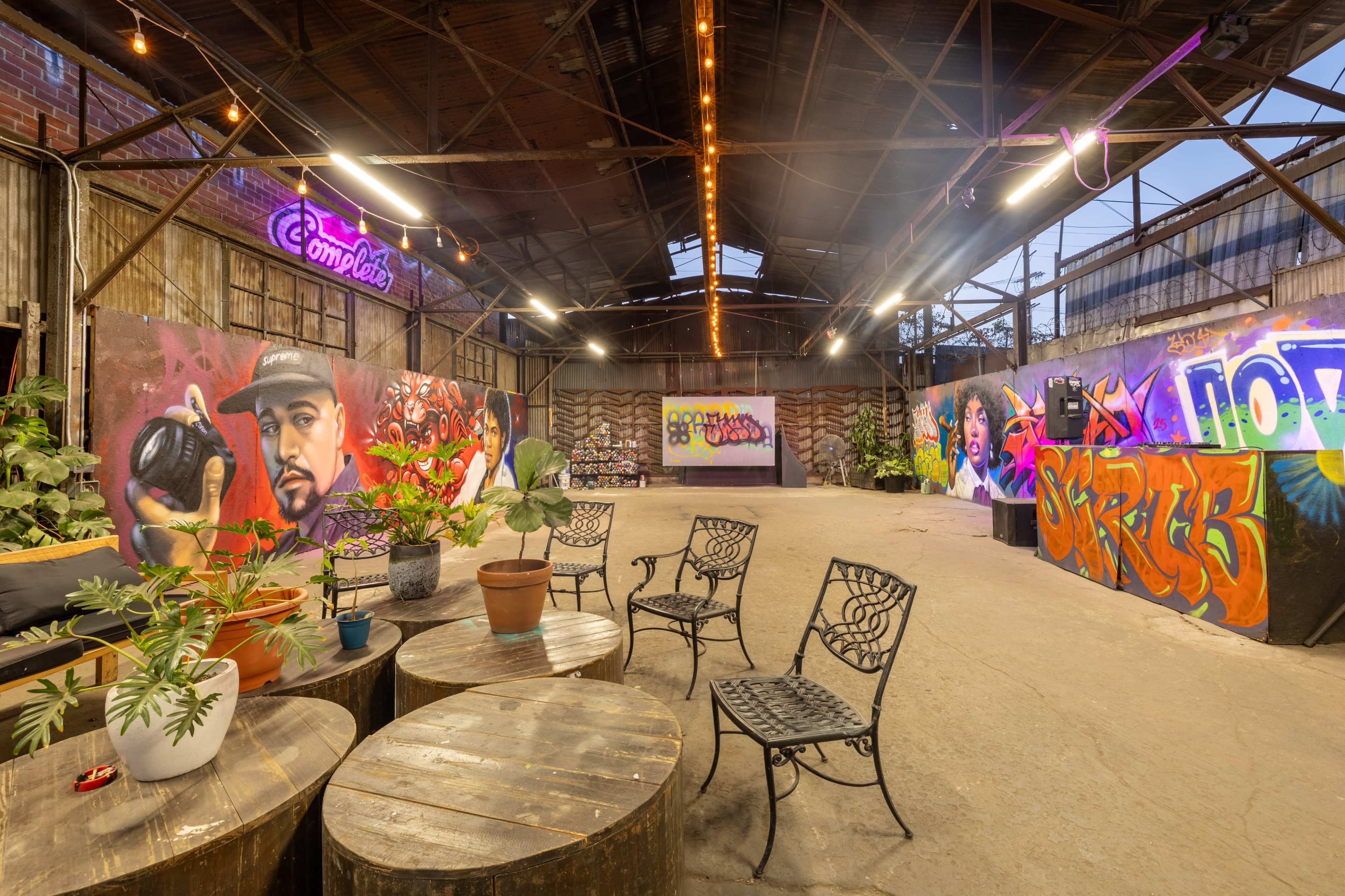 The image shows an industrial-style event space featuring graffiti murals on the walls, plants in pots, and wooden round tables surrounded by metal chairs.