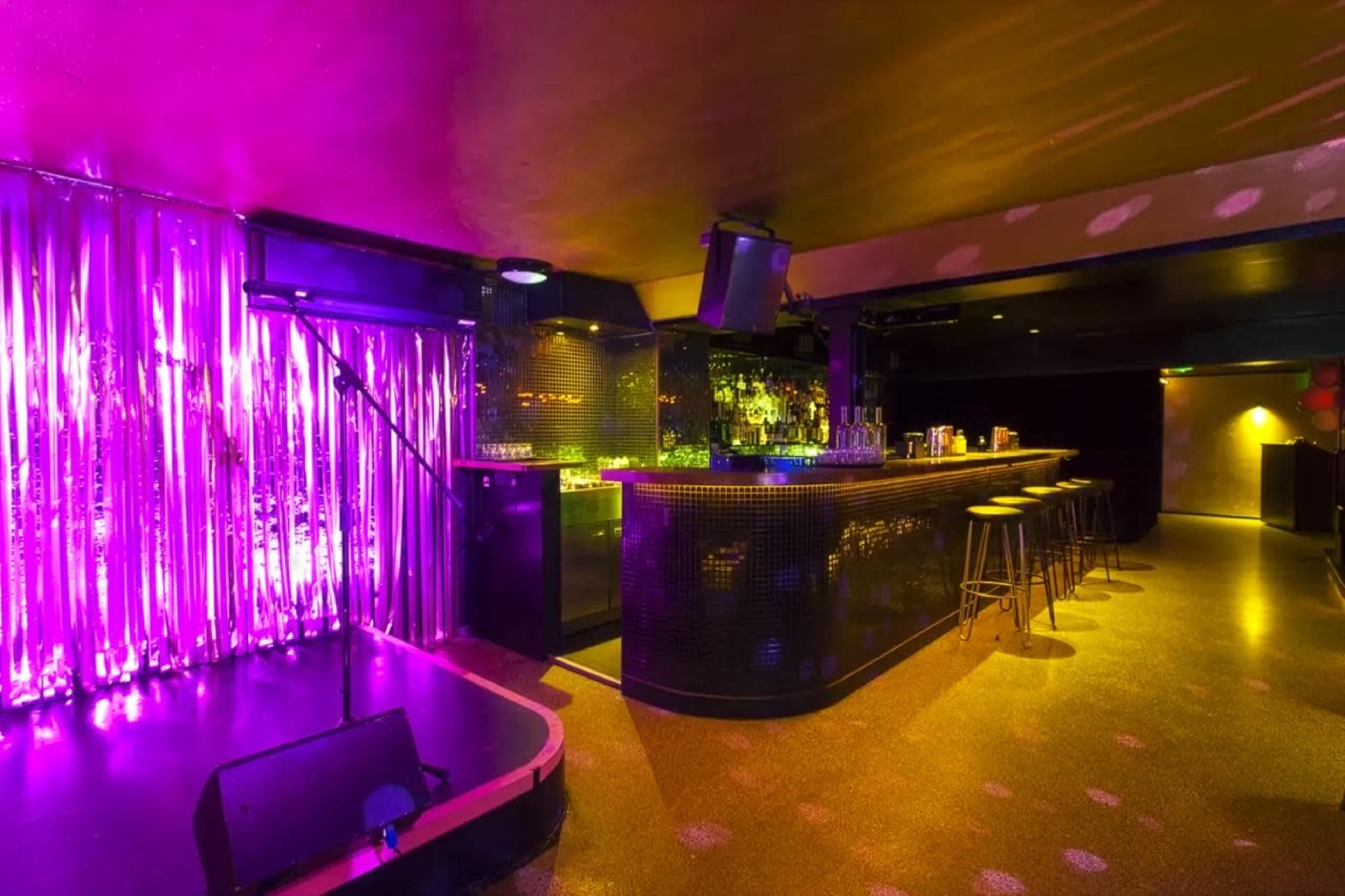 The image shows a dimly lit bar area with a shiny purple backdrop, a long counter with stools, and a small stage with a microphone.