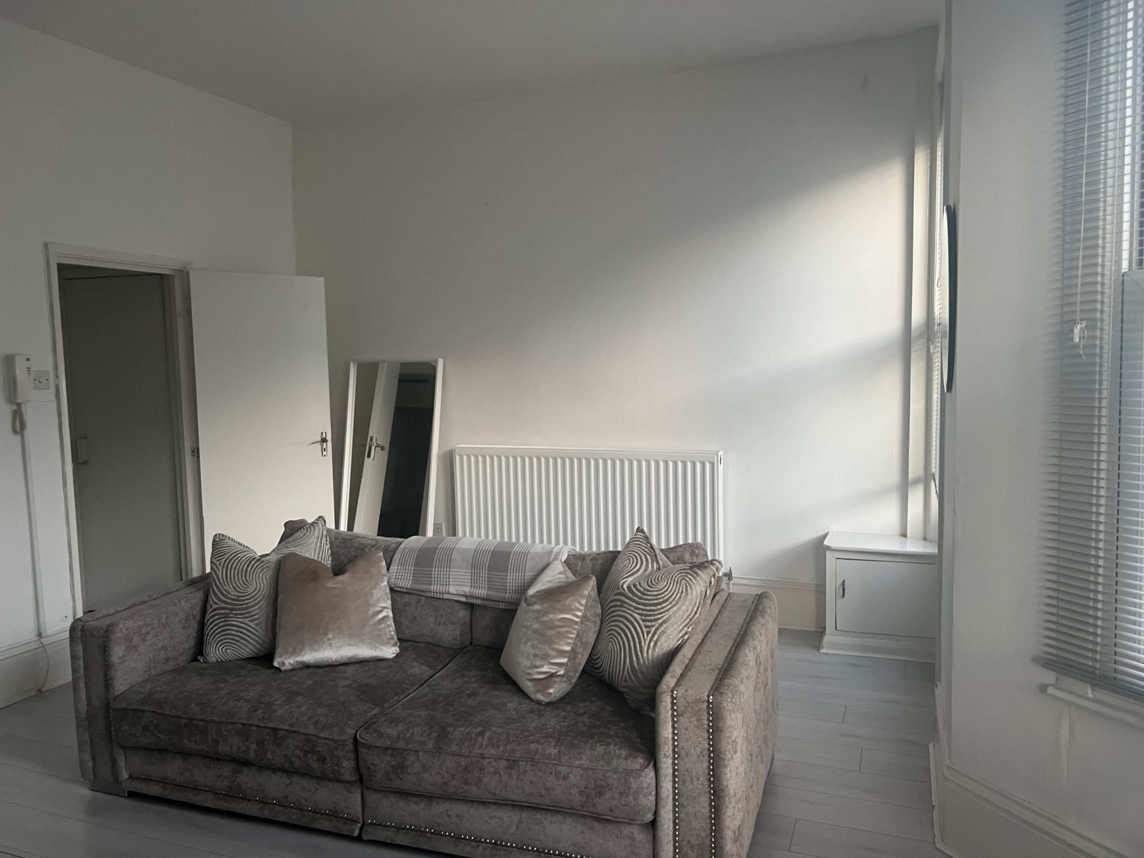 The image shows a minimalist living room with a gray sofa, a large mirror, a radiator, and a small cabinet, all set against white walls and a light wooden floor.
