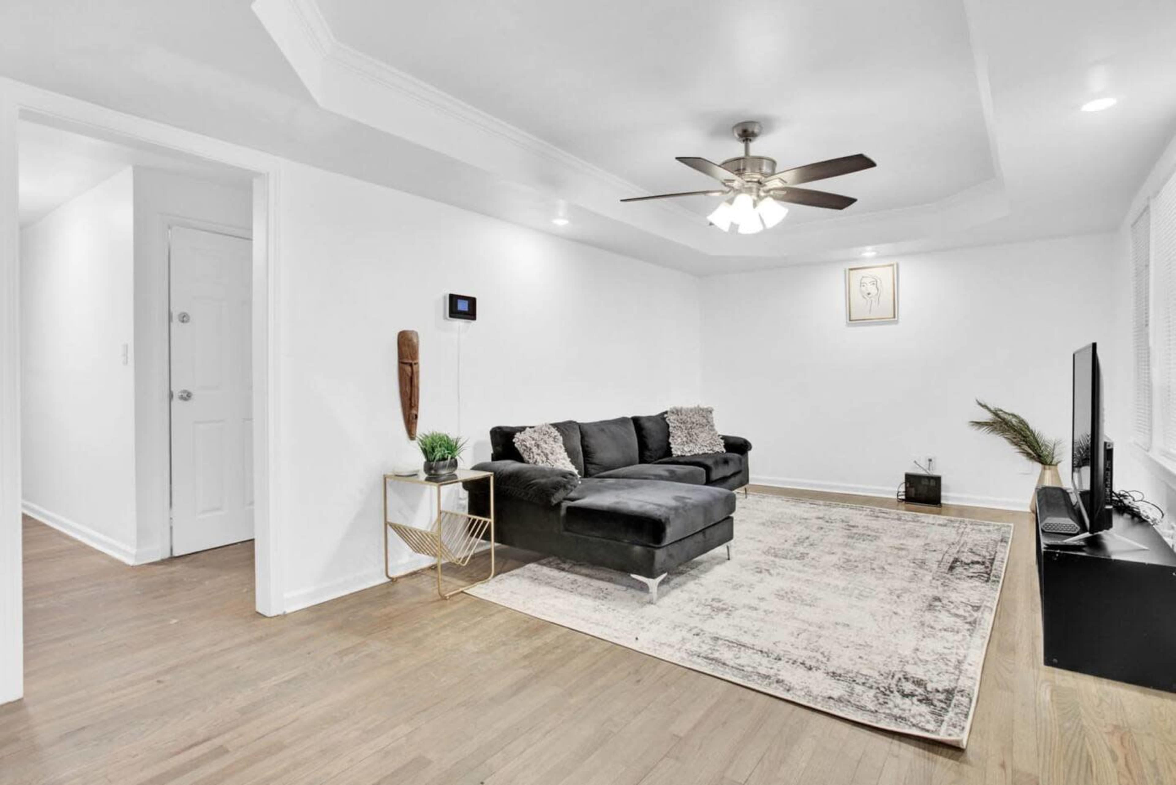 A modern living room with a black sectional sofa, a light area rug, a television, and a ceiling fan.