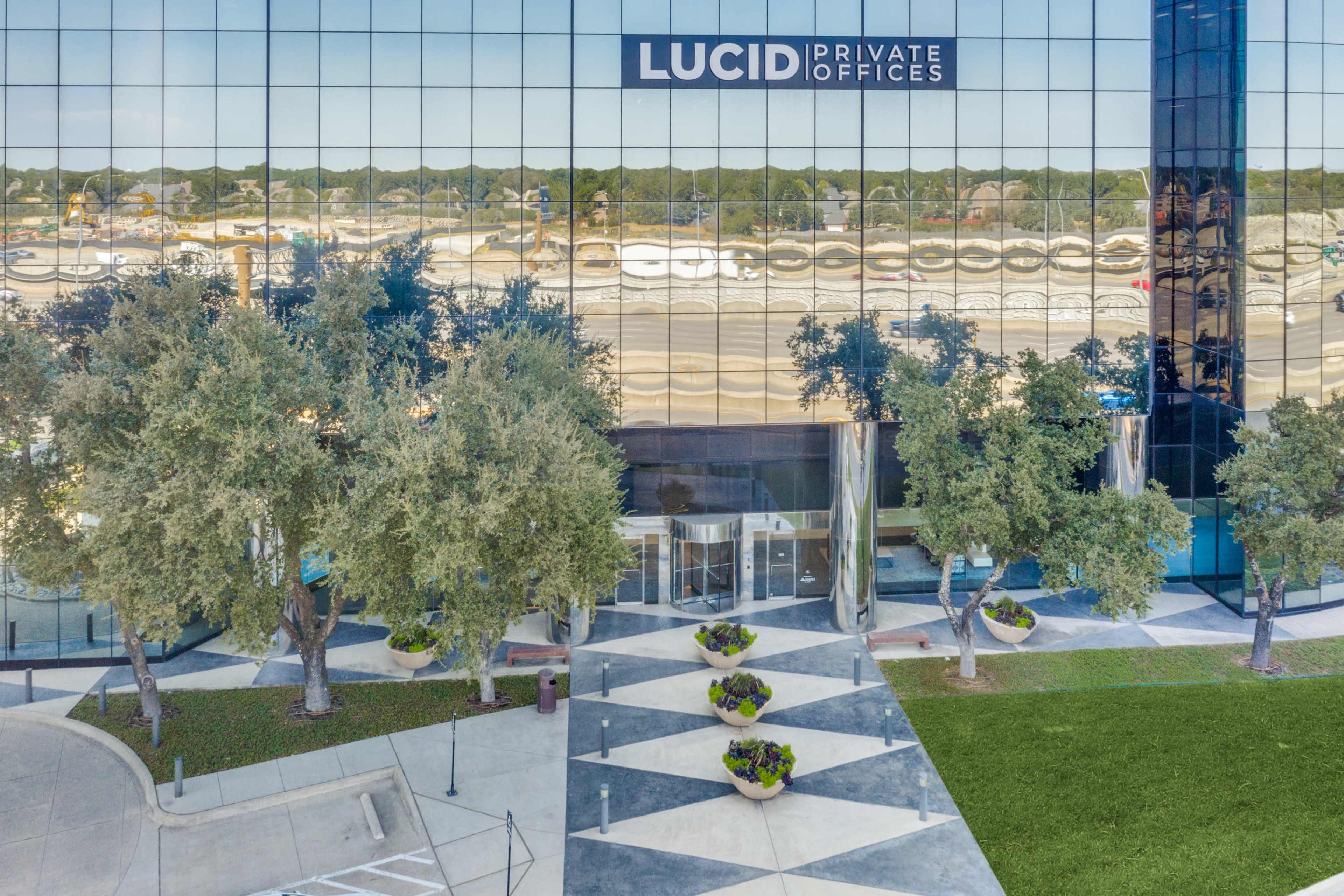 The image shows a modern office building with reflective glass windows, featuring the signage "LUCID PRIVATE OFFICES" above the entrance, surrounded by landscaped trees and planters.