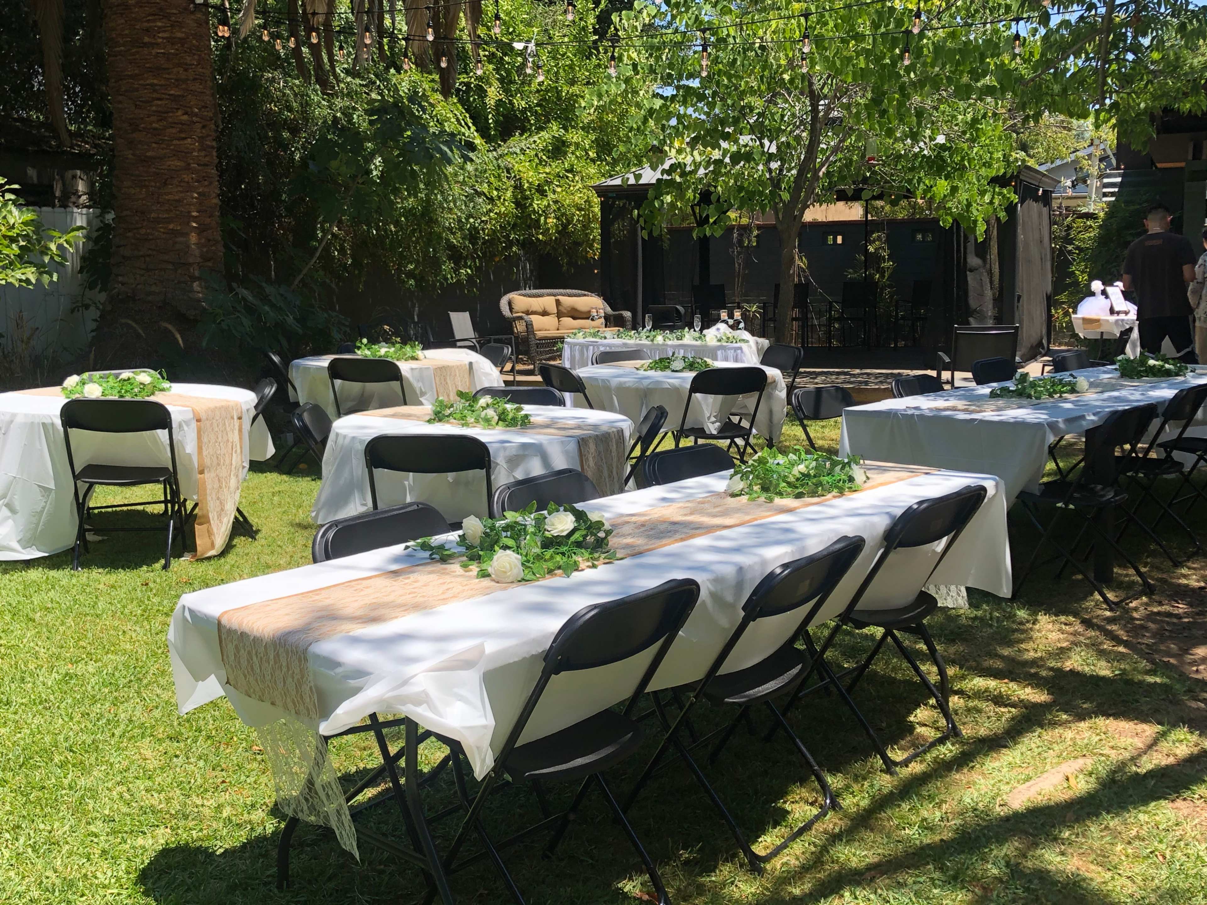 A green outdoor space set up for an event, featuring several long tables adorned with tablecloths and floral centerpieces.