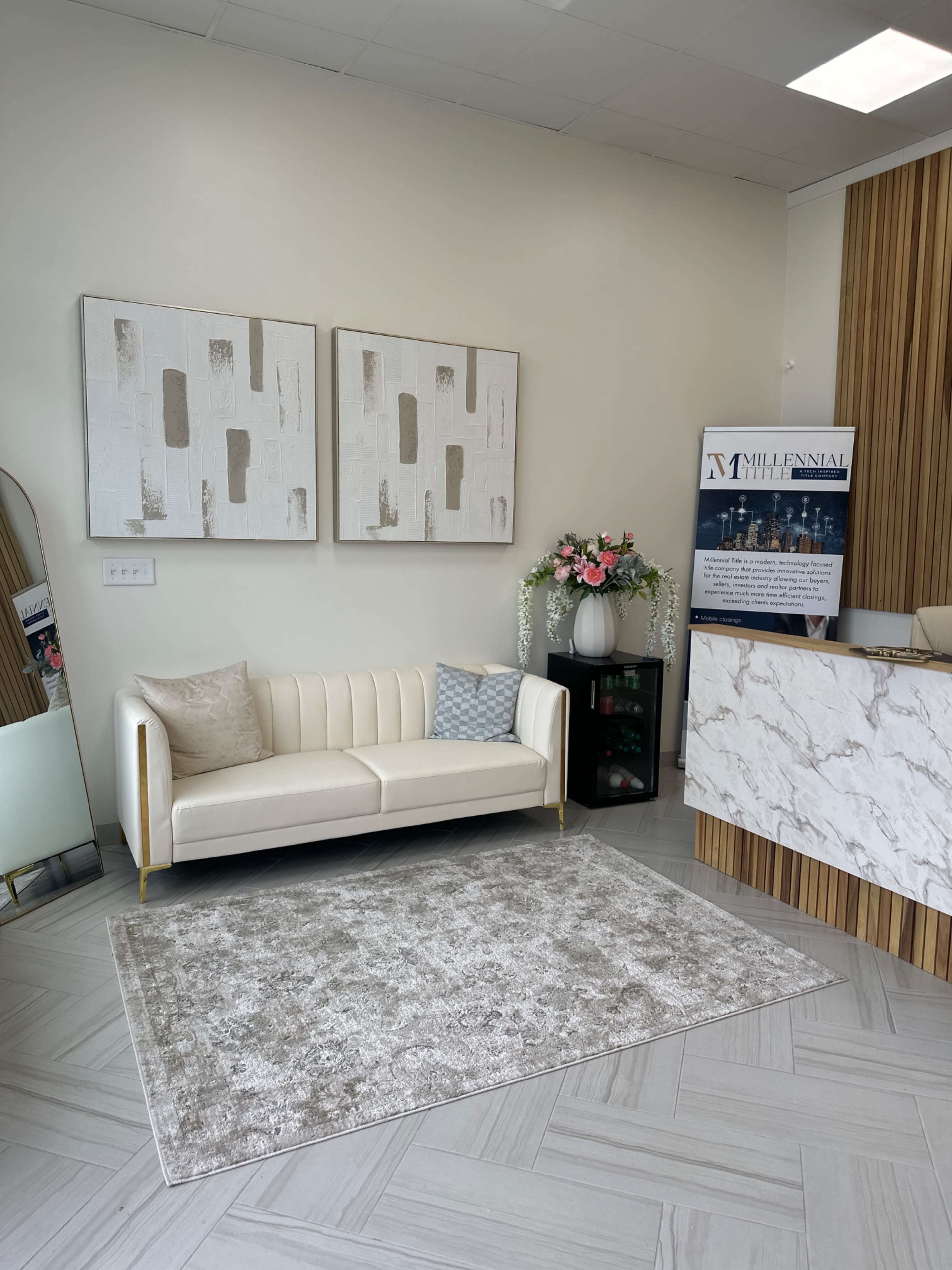 The image shows a modern reception area featuring a white leather sofa, decorative wall art, a gray rug, and a stylish reception desk with floral arrangements.