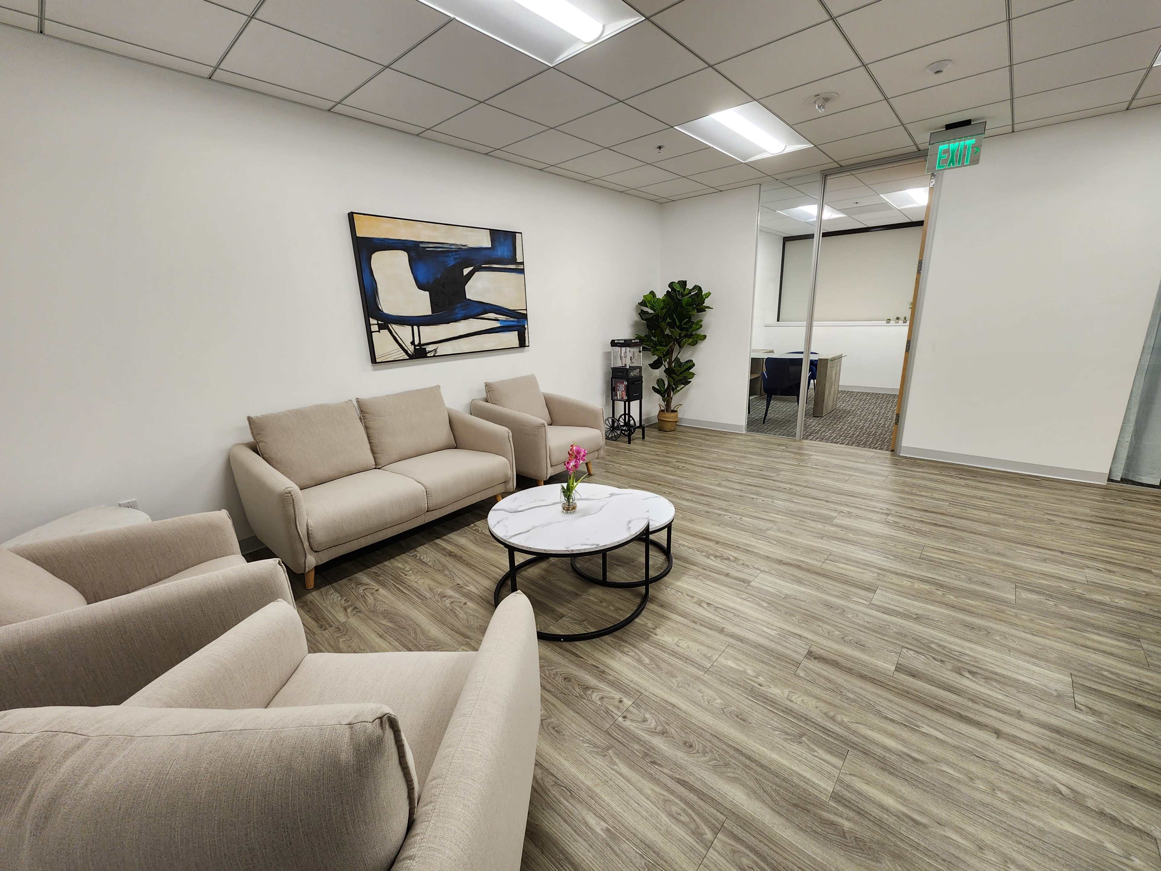 The image shows a modern waiting area with two light-colored sofas, a round coffee table, a decorative plant, and an abstract painting on the wall.