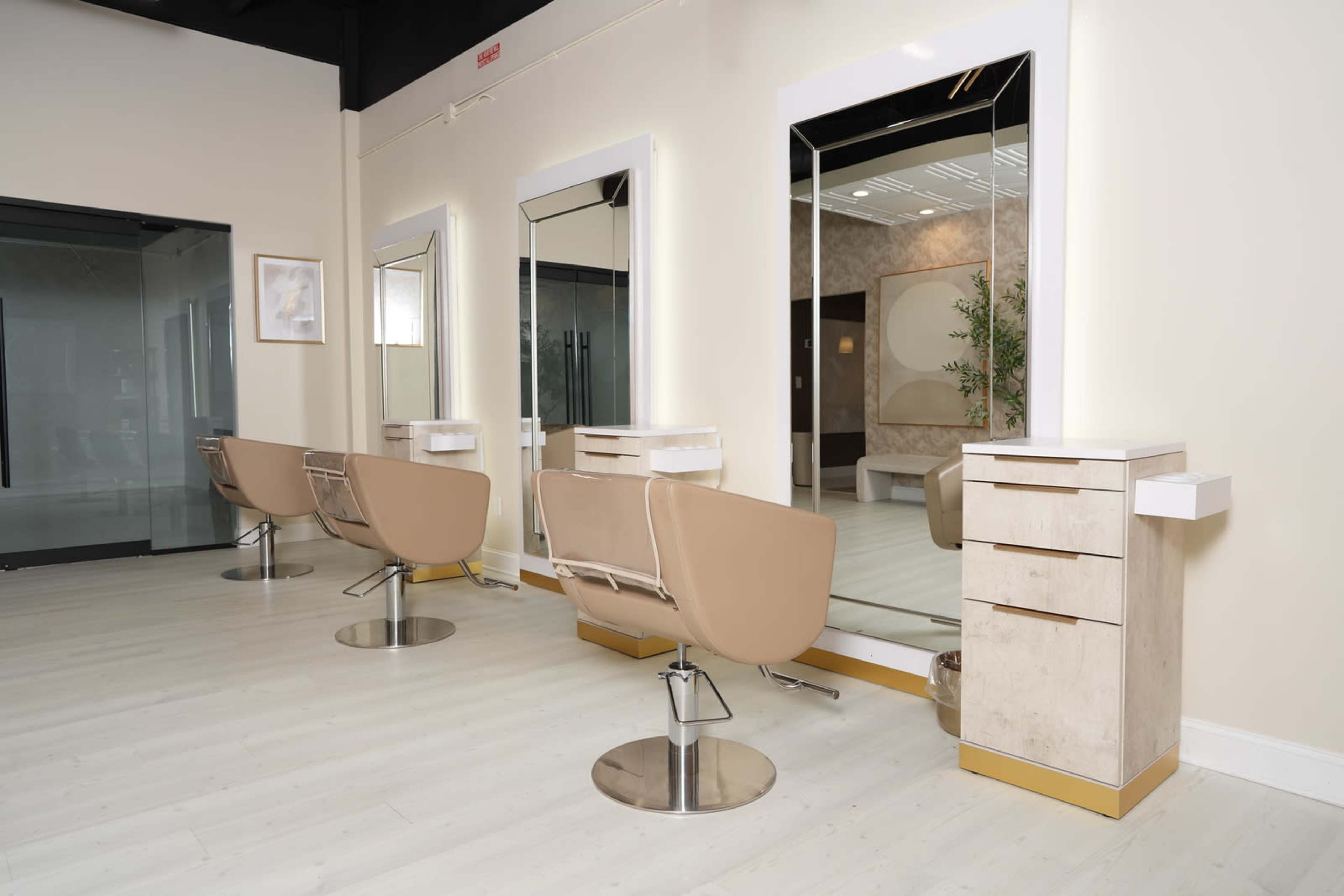 A modern salon interior featuring three styling stations with mirrors and beige chairs, set against light wooden flooring.