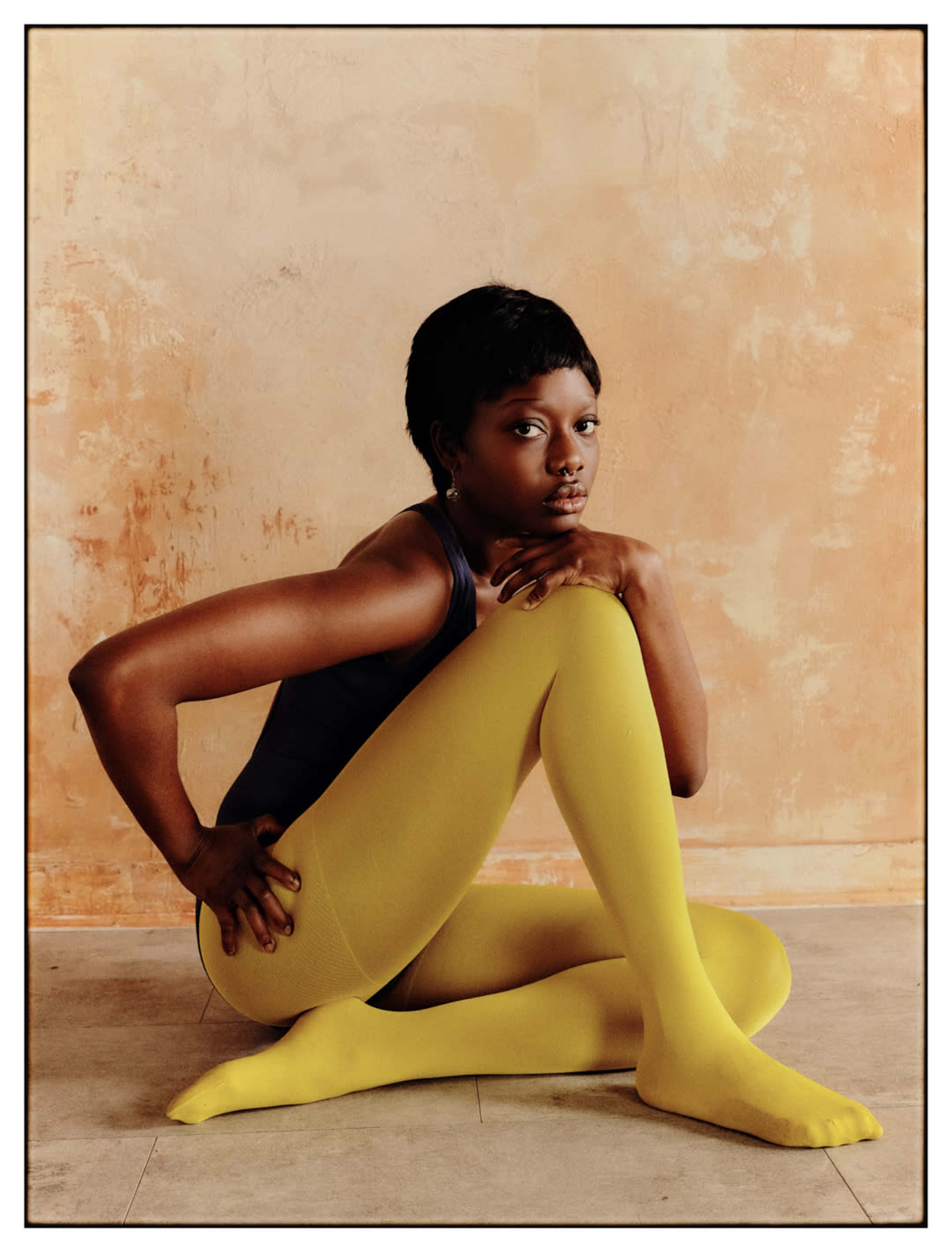 A person sits on the floor with their legs crossed, wearing a dark top and bright yellow tights, against a textured beige wall.