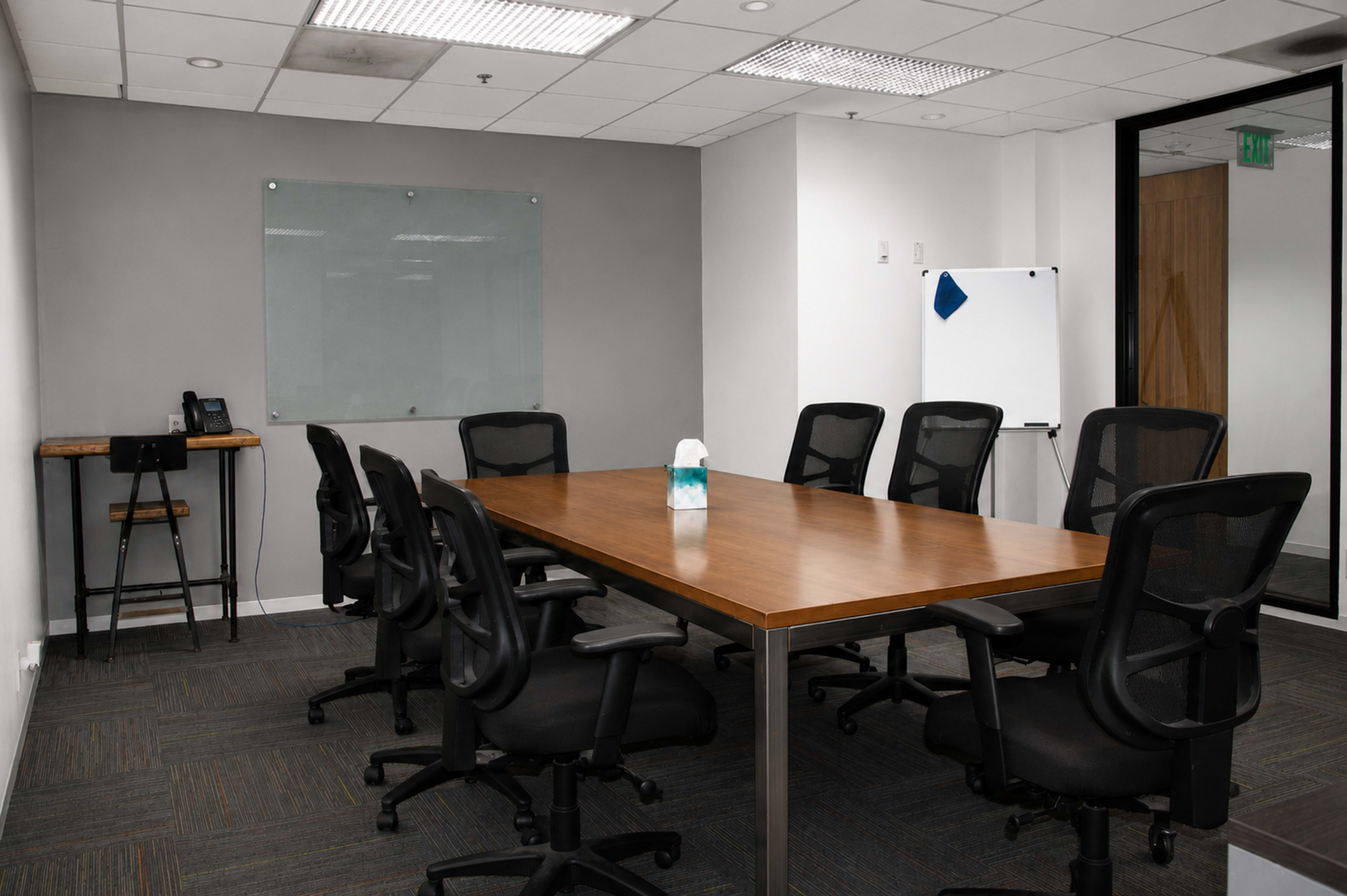 The image shows a modern conference room furnished with a large wooden table, several black rolling chairs, a whiteboard, and a glass partition.