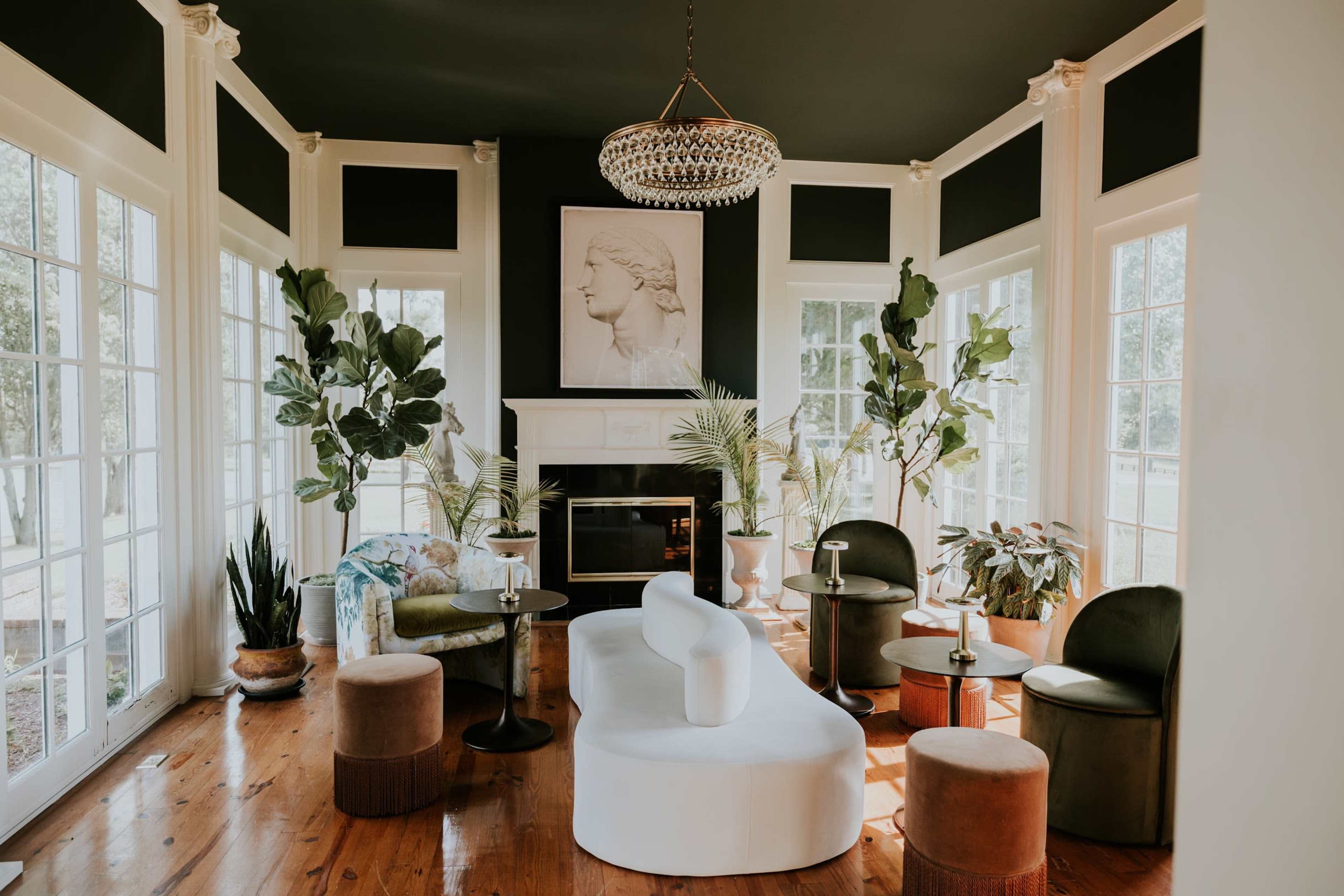 The room features a modern seating area with a white couch, green and brown chairs, potted plants, and large windows allowing natural light.