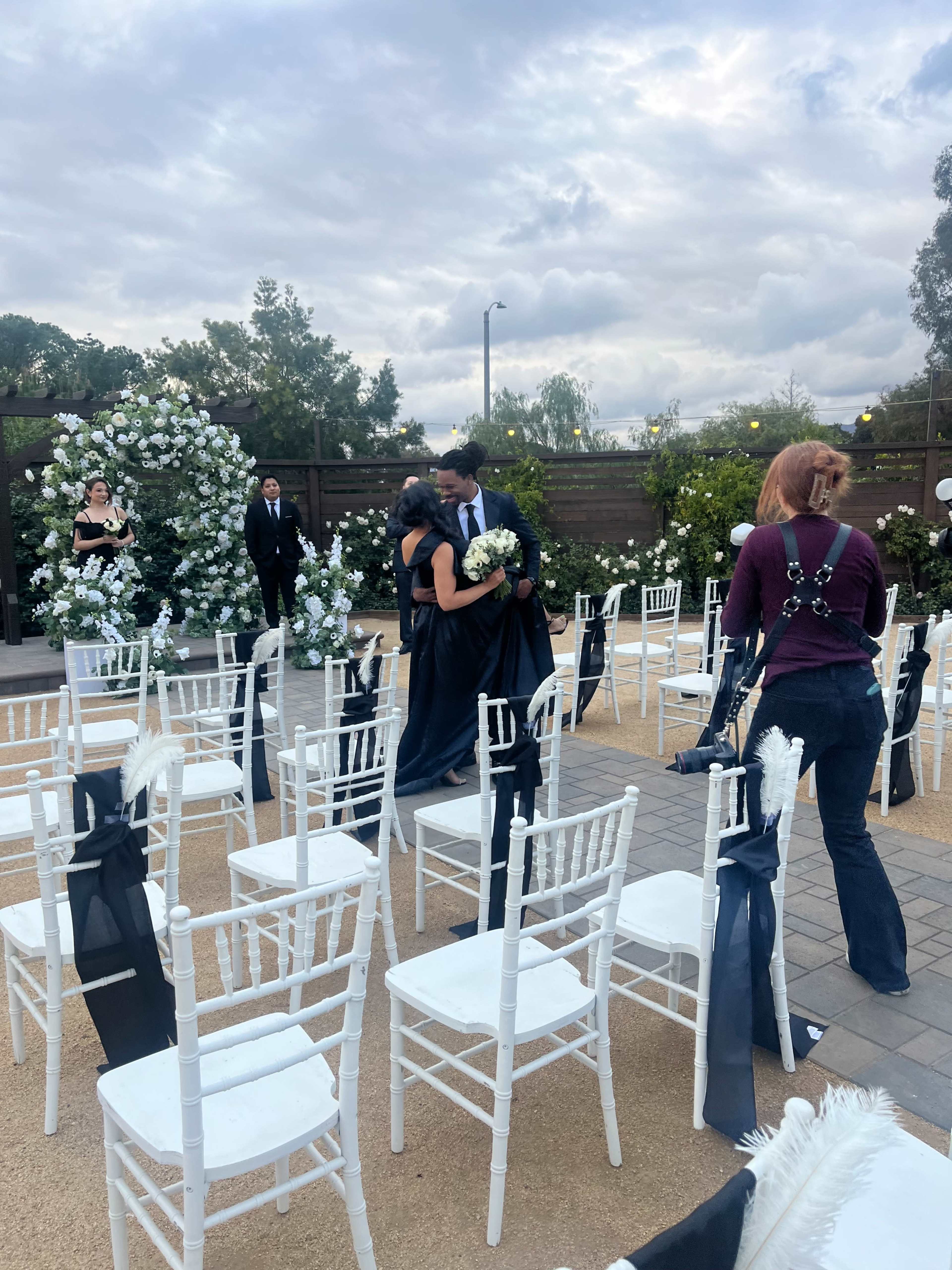 A bride and groom are seen walking among white chairs decorated with black sashes in an outdoor wedding venue.