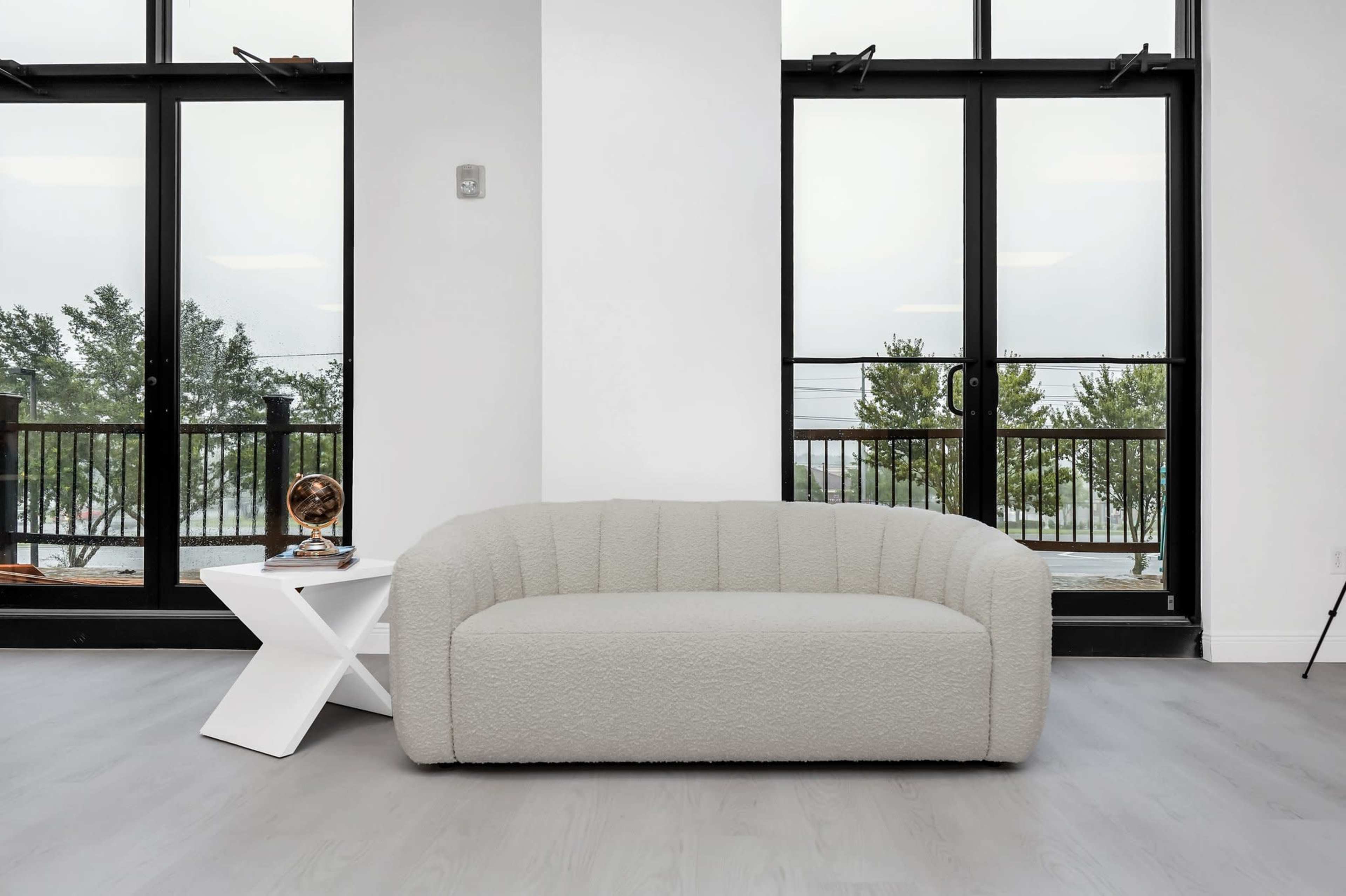 A light-colored, upholstered sofa is placed next to a white angular side table in front of large glass doors overlooking a balcony and trees.