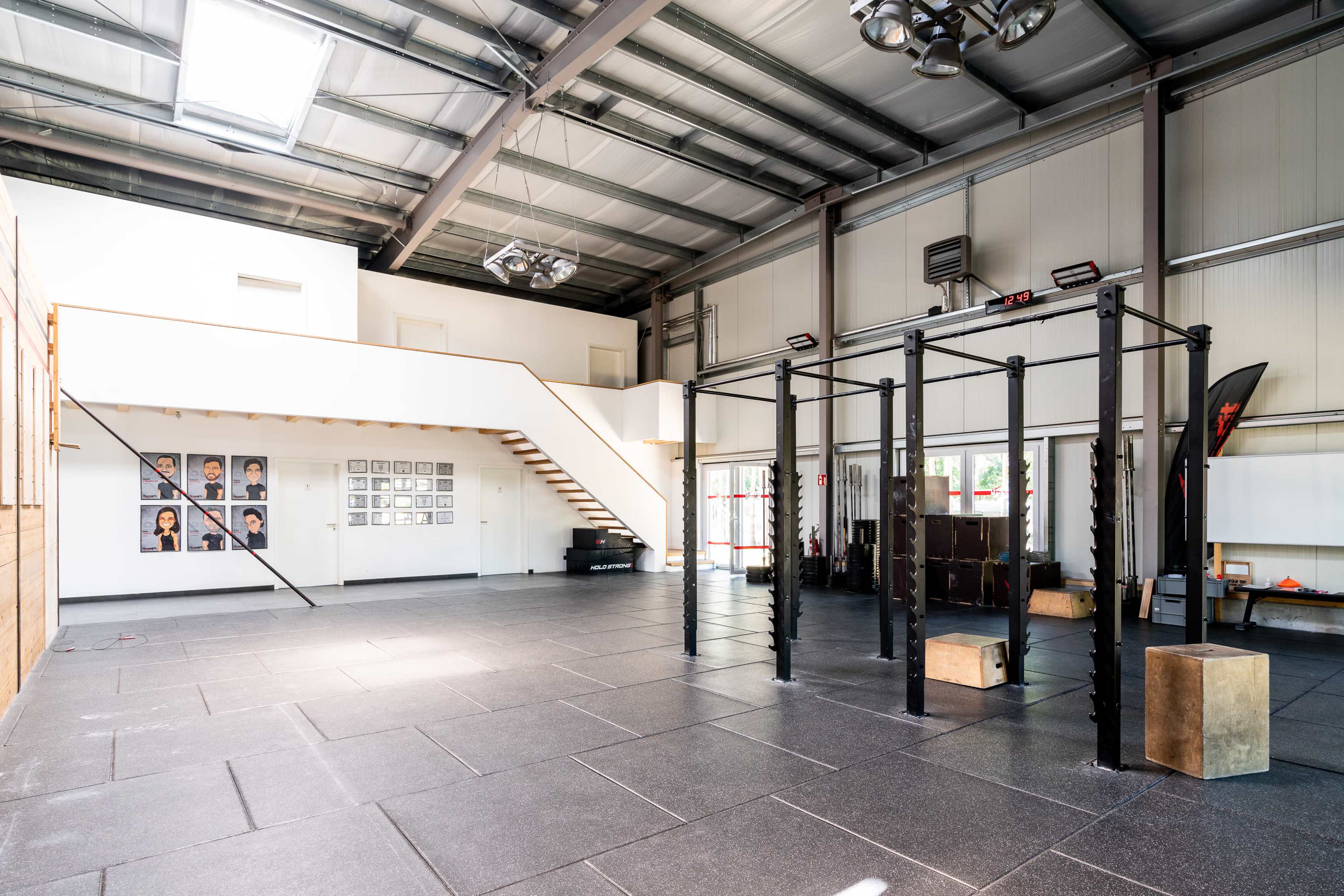 An empty gym interior featuring a pull-up bar setup, a staircase, and framed photographs on the wall.