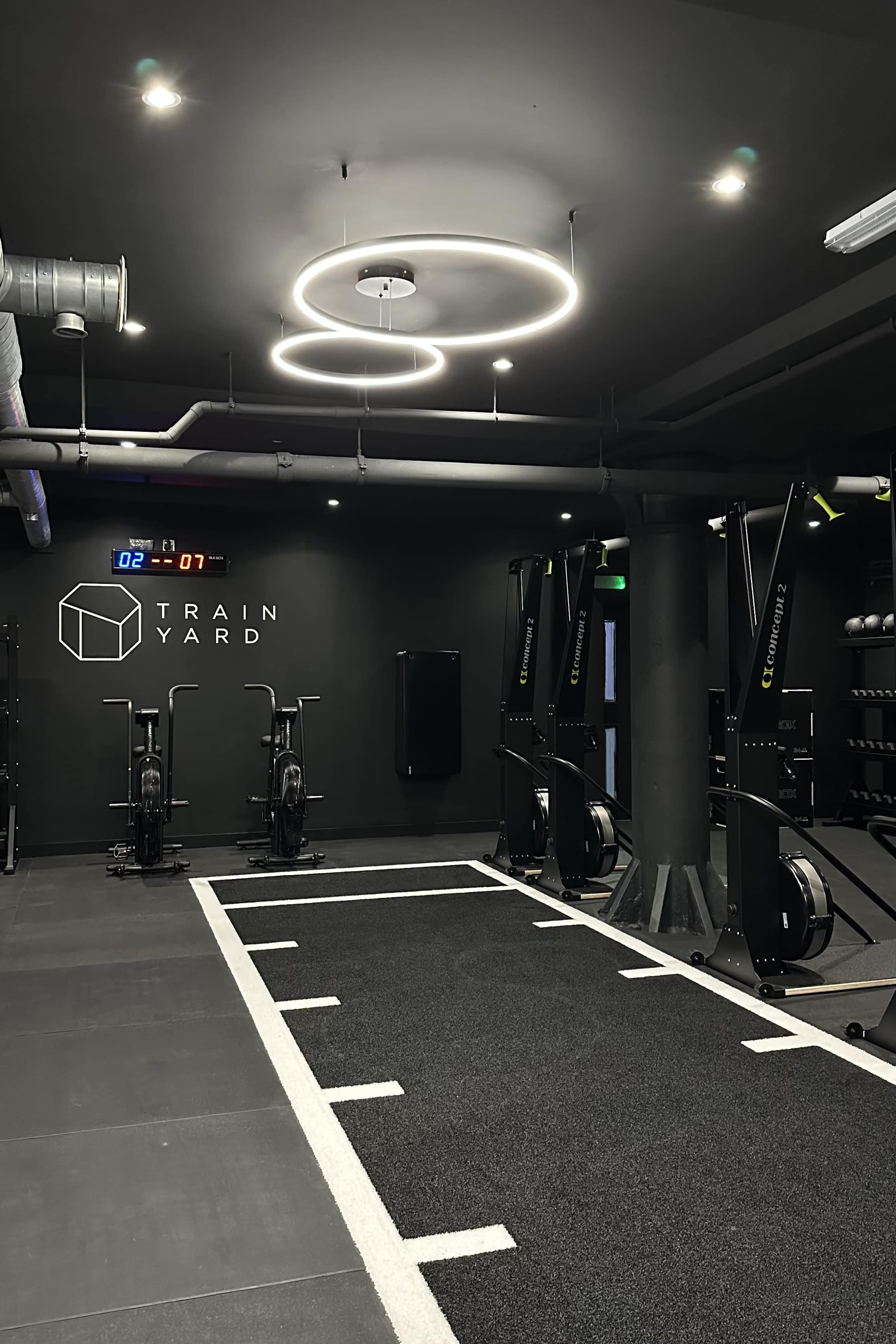 The image shows a modern indoor gym with exercise bikes, a black turf area, and circular lighting fixtures.