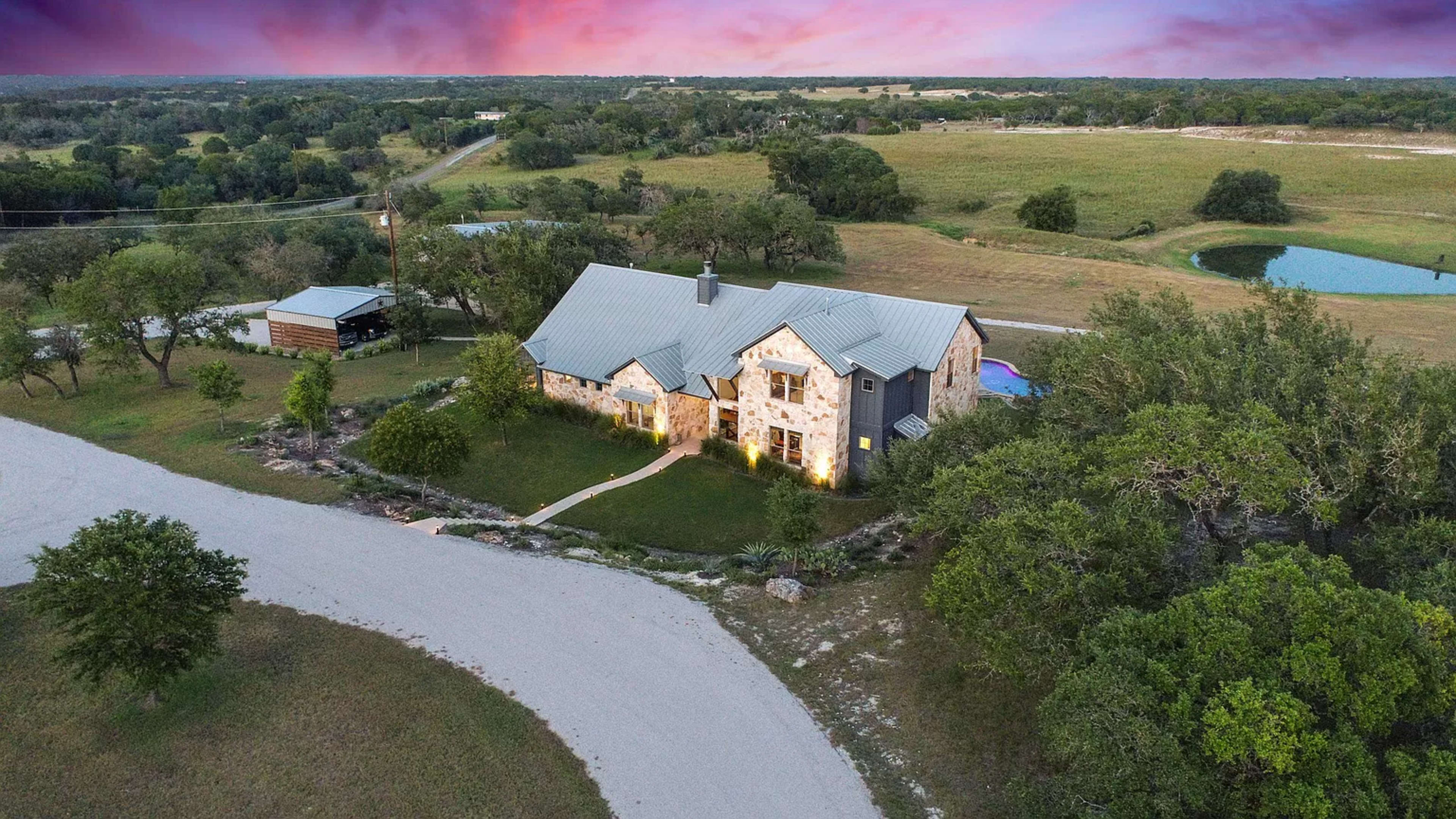 The image shows a large stone house with a metal roof, surrounded by green fields and trees, situated at a crossroads with a gravel road in the foreground.