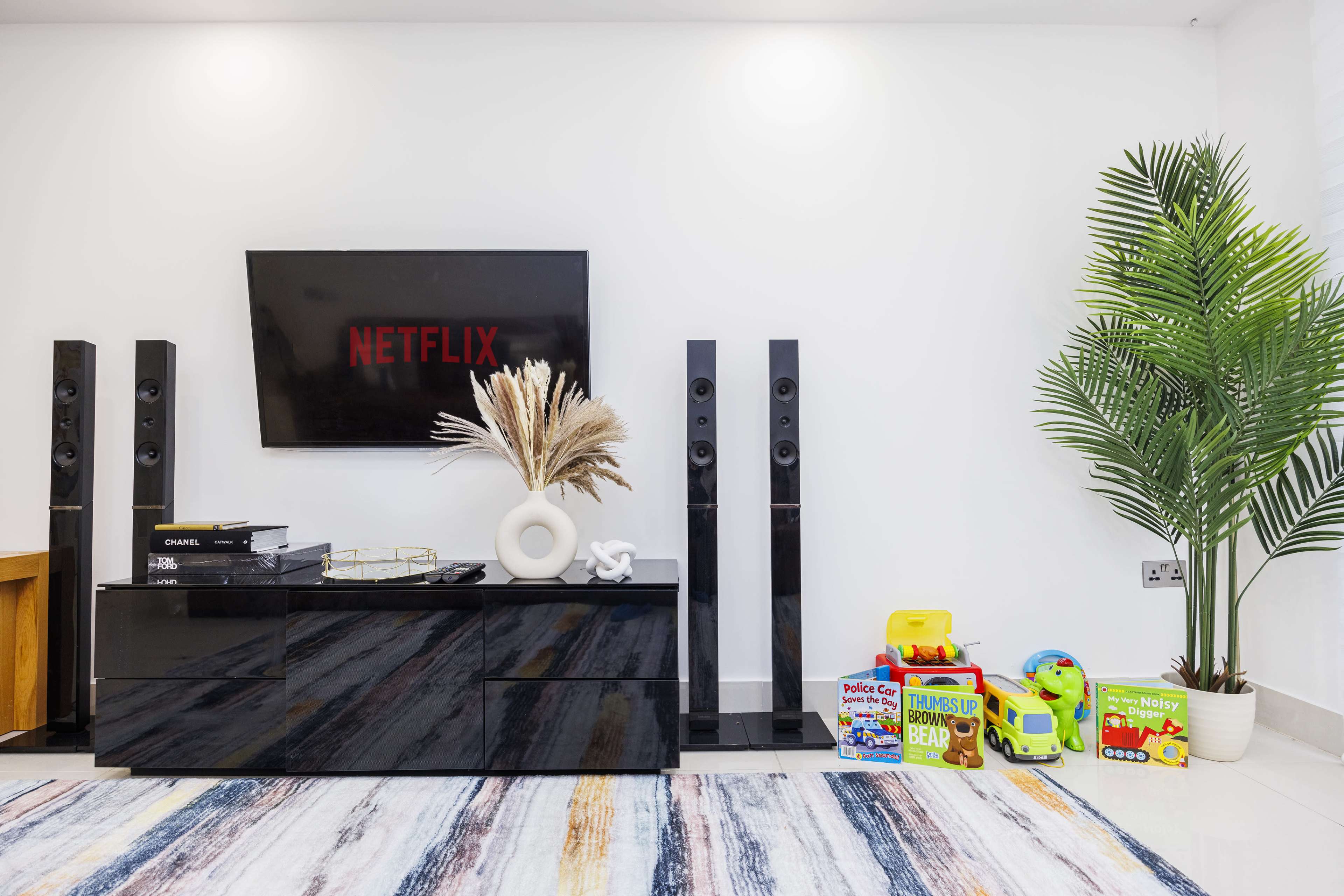 A modern living room features a black entertainment unit with a Netflix display, flanked by tall speakers, while colorful children's toys are arranged beside a potted plant.