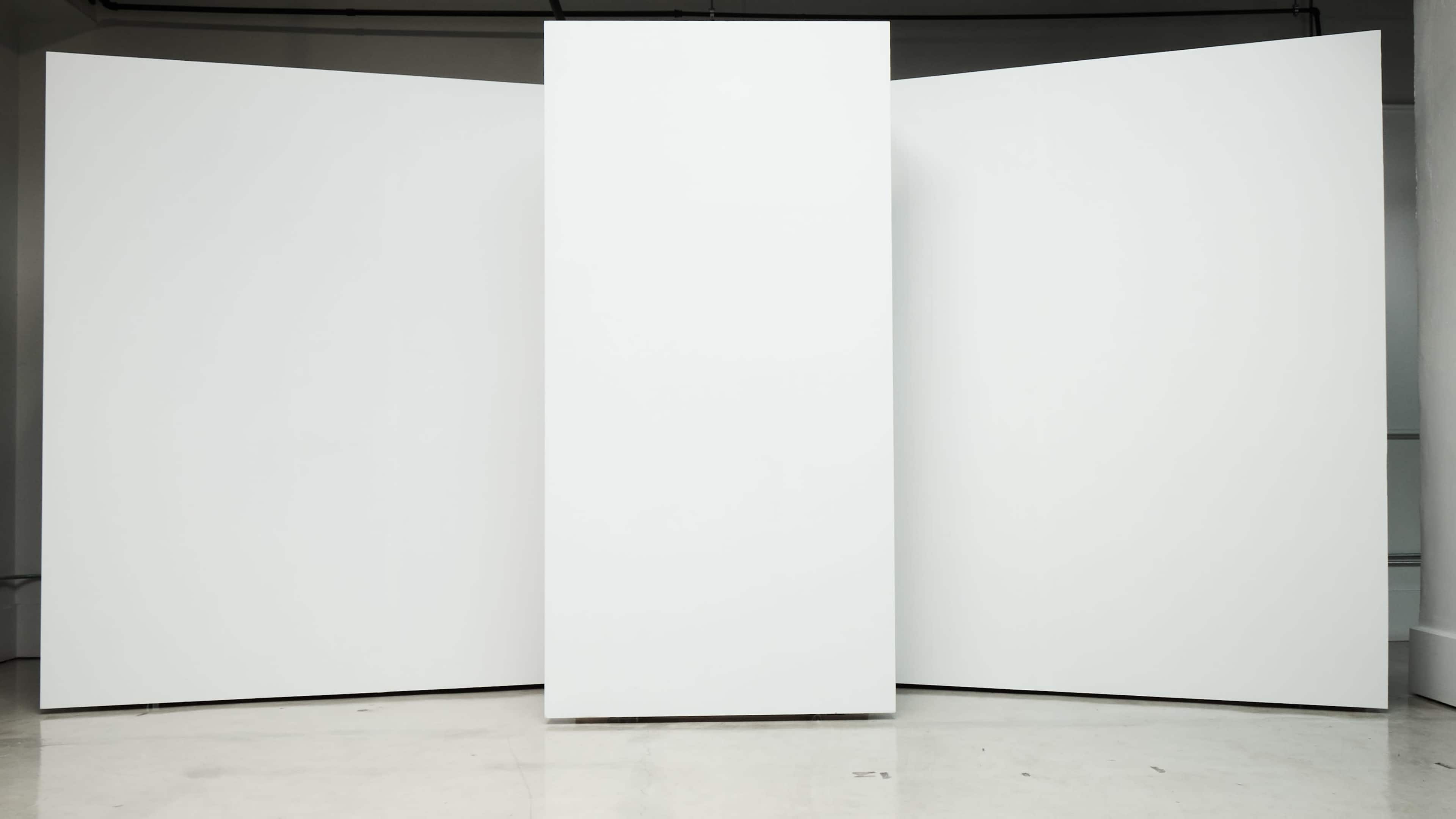The image shows three large, blank white panels arranged in a triangular formation in a minimalistic space.