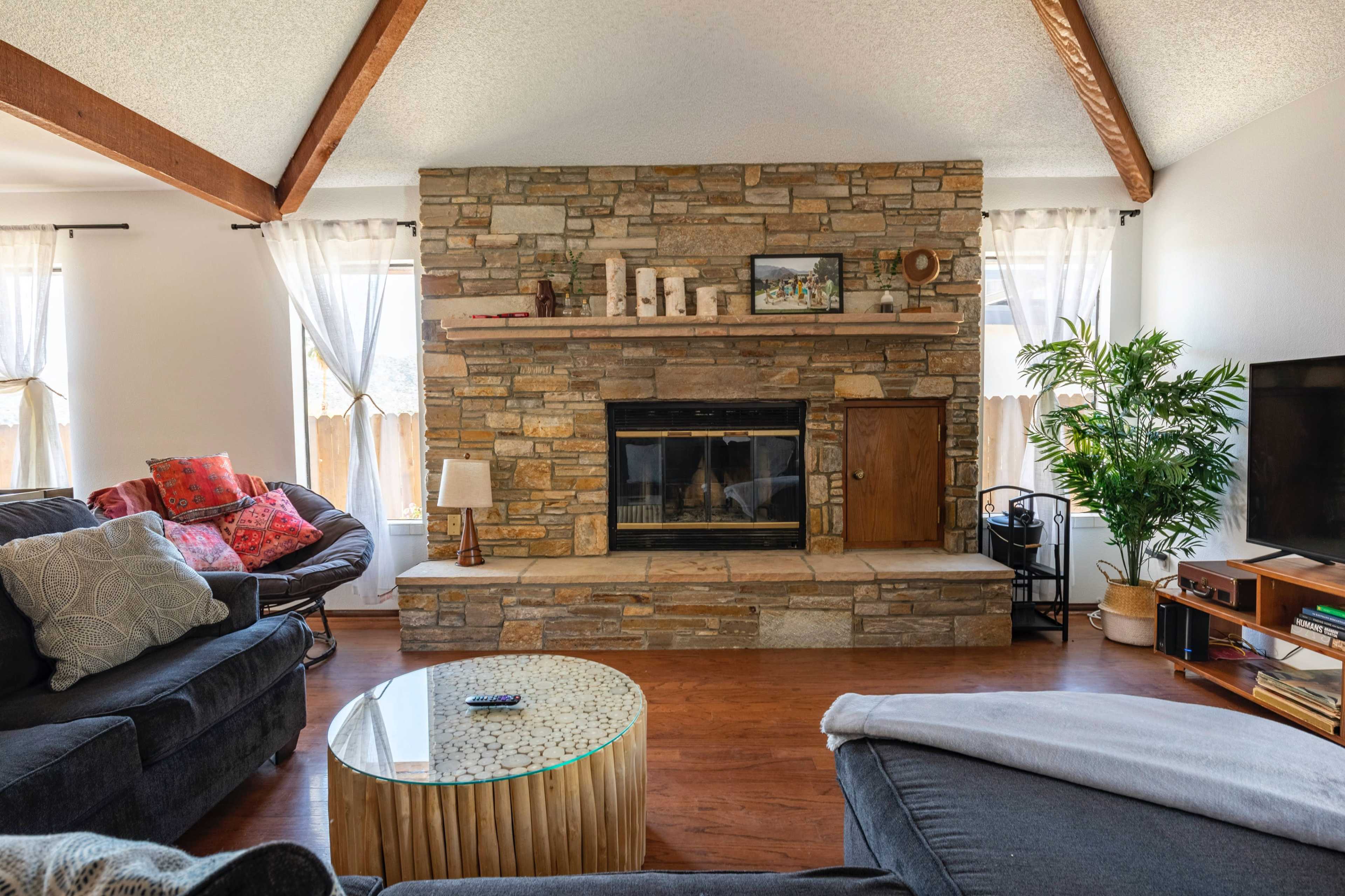 A living room with a stone fireplace, two sofas, a round coffee table, and large windows dressed in sheer curtains.