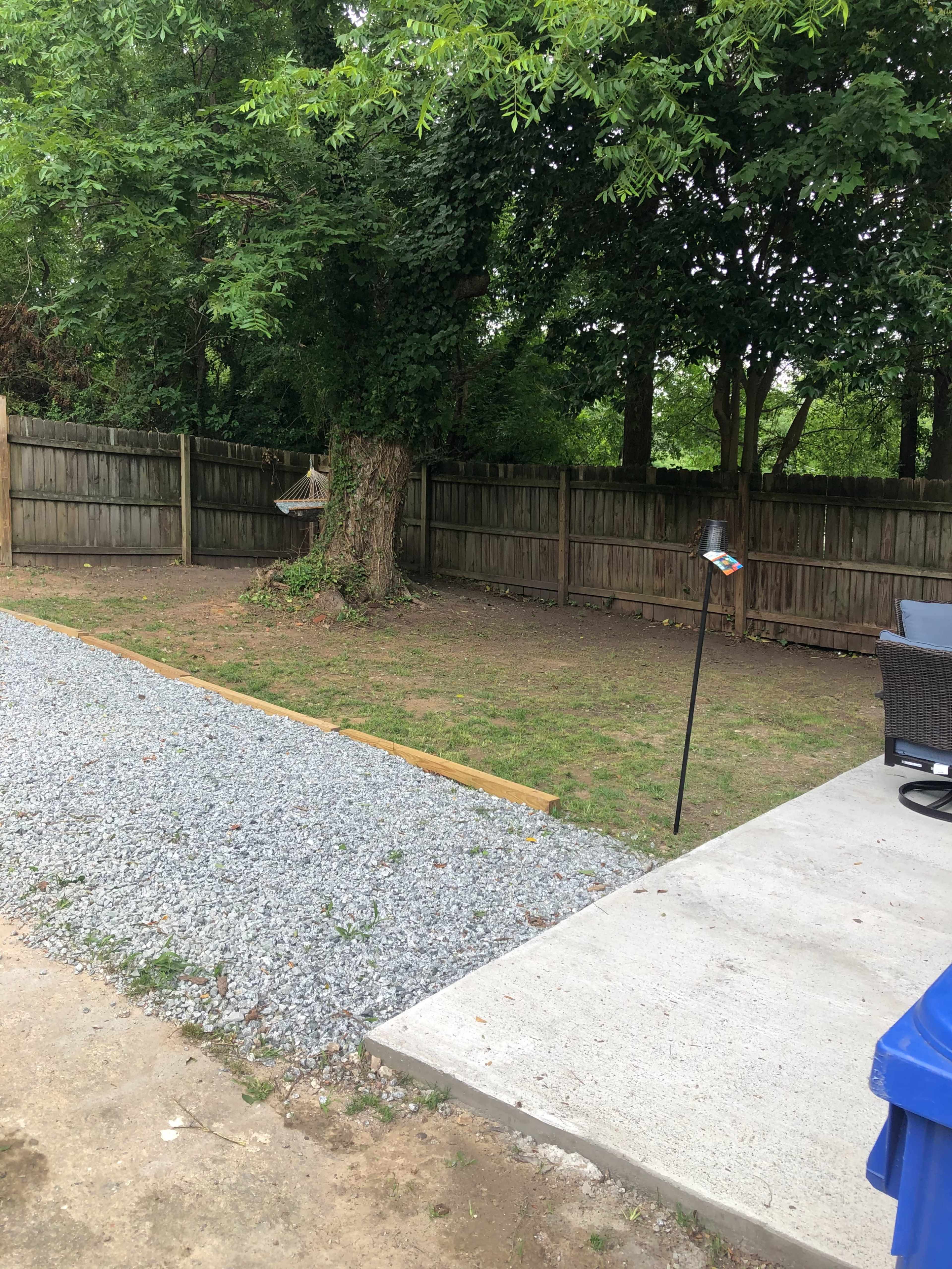 A gravel walkway leading to a grassy area enclosed by a wooden fence, with a cement patio and outdoor furniture on one side.