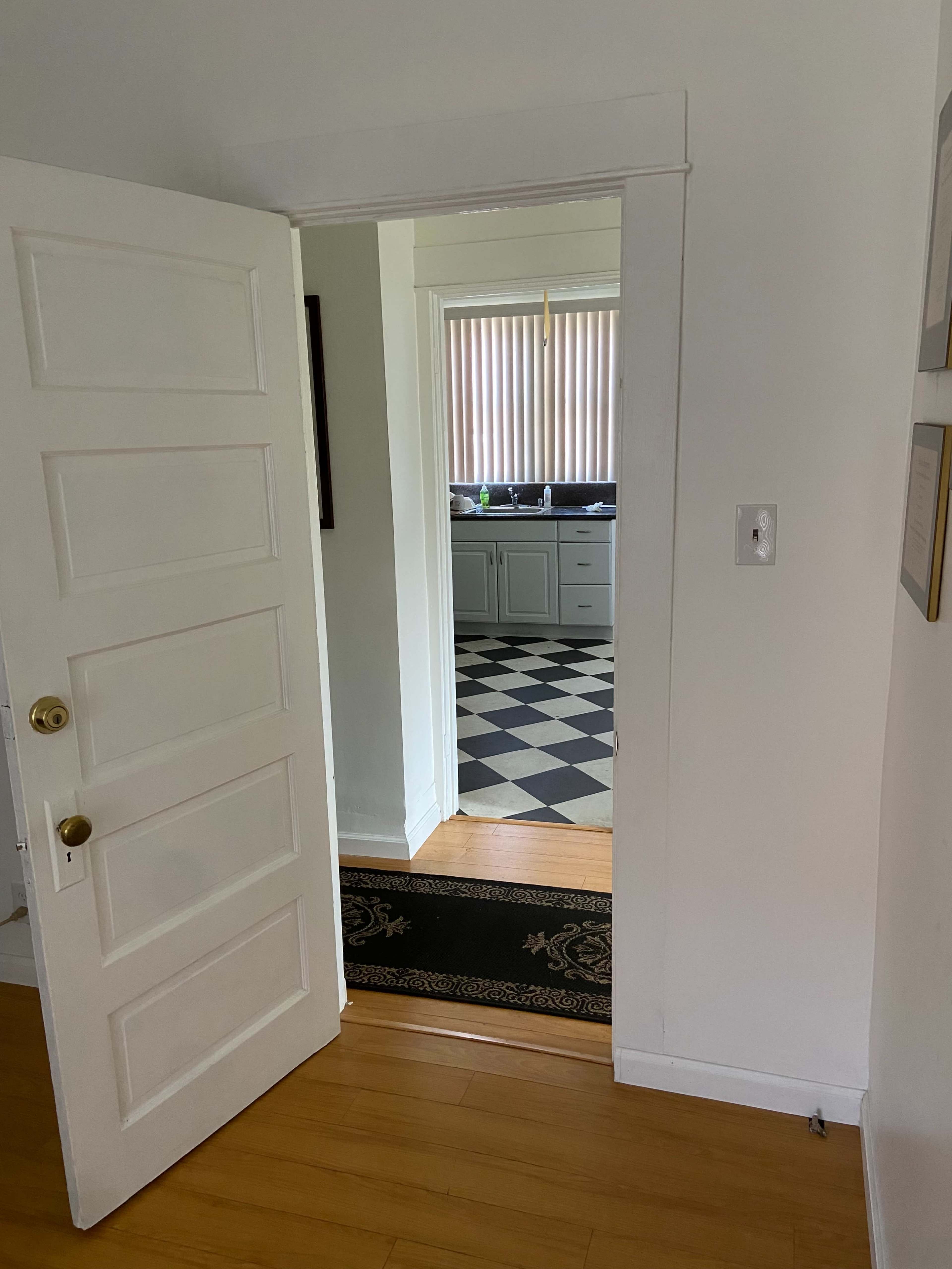 An open door reveals a view of a kitchen with checkered flooring and natural light filtering through window blinds.