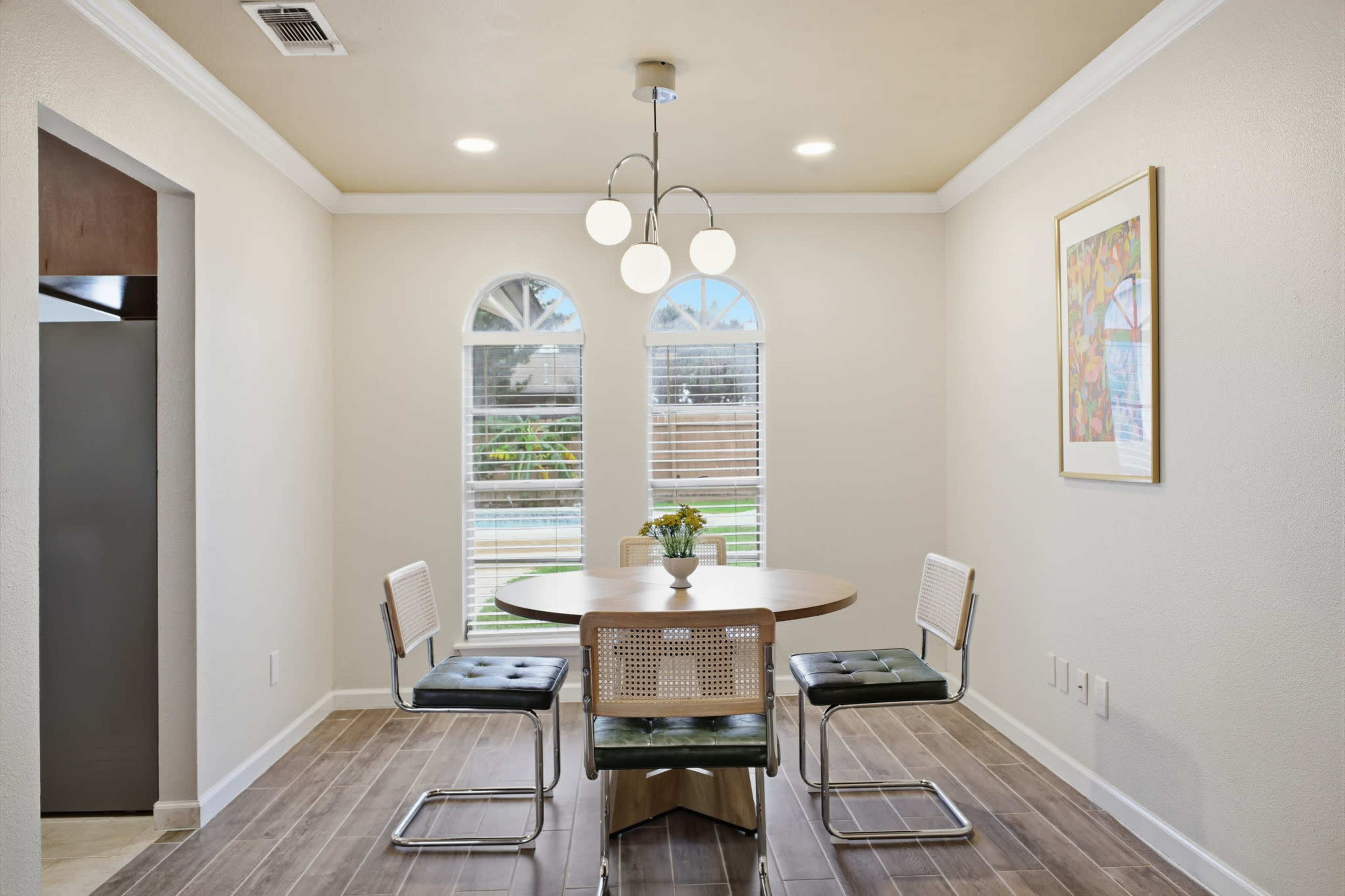 The image shows a small dining area featuring a round wooden table with four chairs and large windows providing natural light.