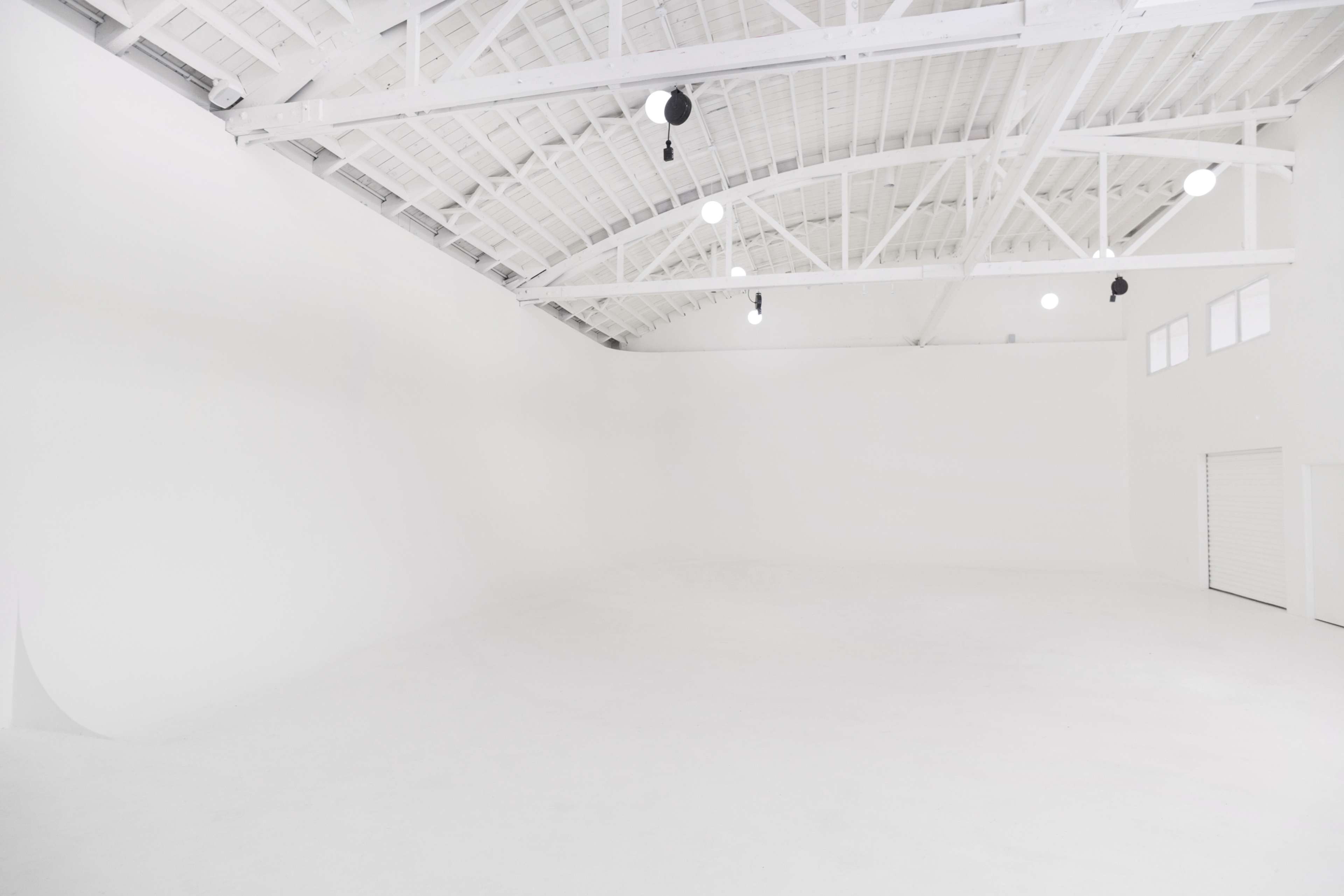A spacious, empty studio with white walls and a high, ceiling featuring exposed beams and hanging lights.