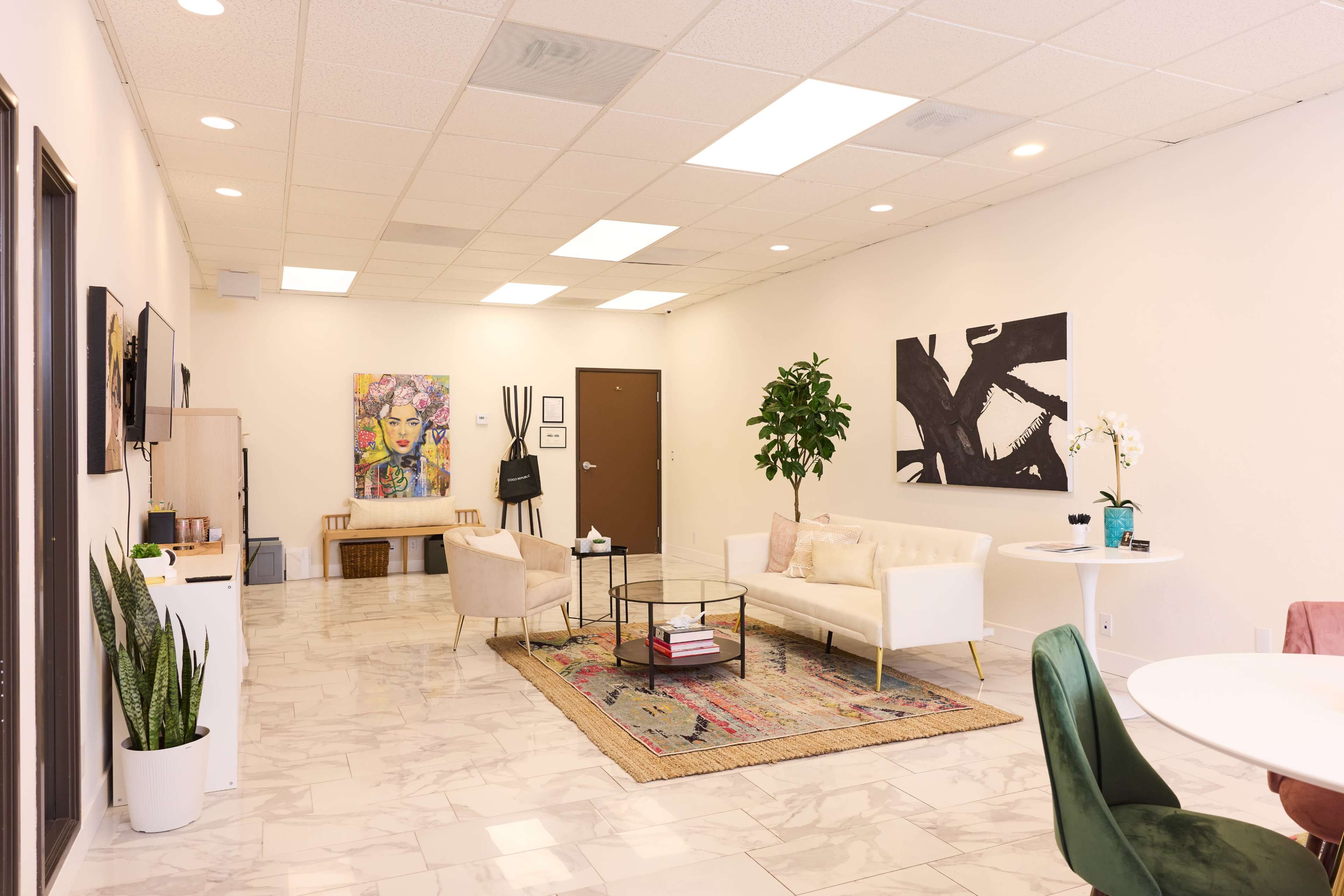 A spacious waiting area with light-colored walls, a large rug on marble flooring, two sofas, a table with chairs, and a plant, along with artwork displayed on the walls.