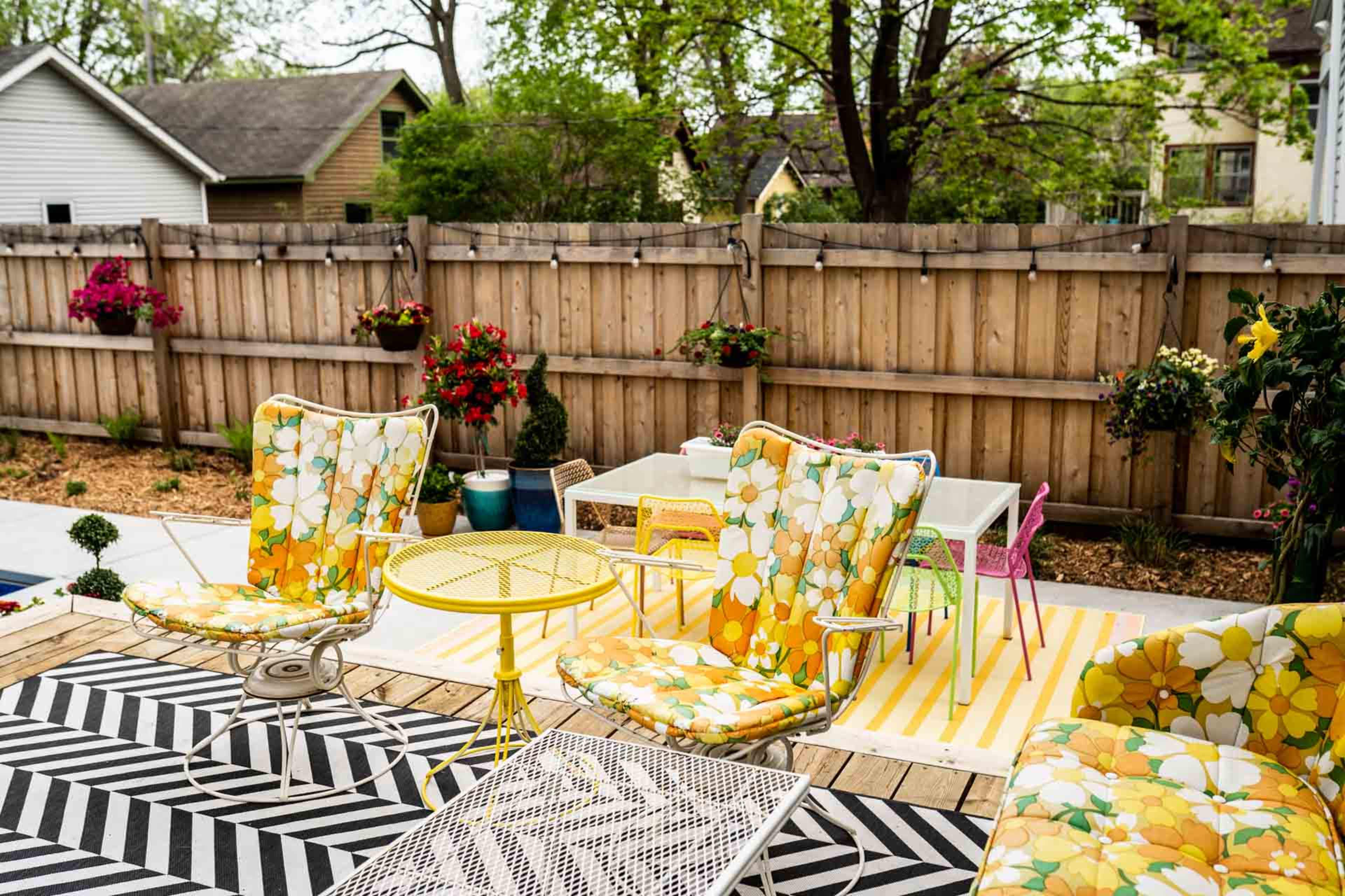 A backyard patio with floral-patterned chairs, a small yellow table, and a striped rug, surrounded by a wooden fence and potted plants.