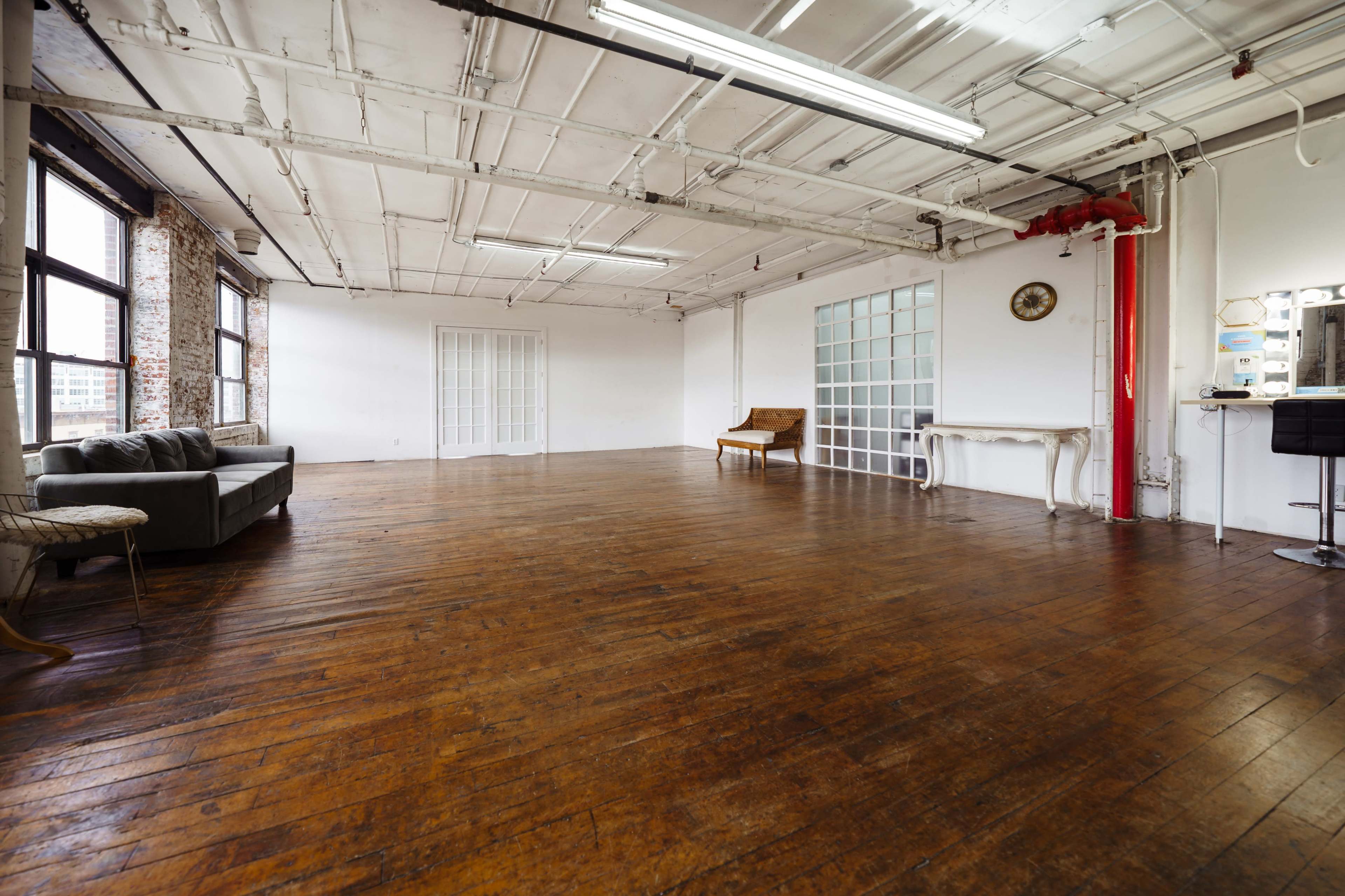 The image shows a spacious, empty room with wooden floors, large windows, and minimalist furniture.