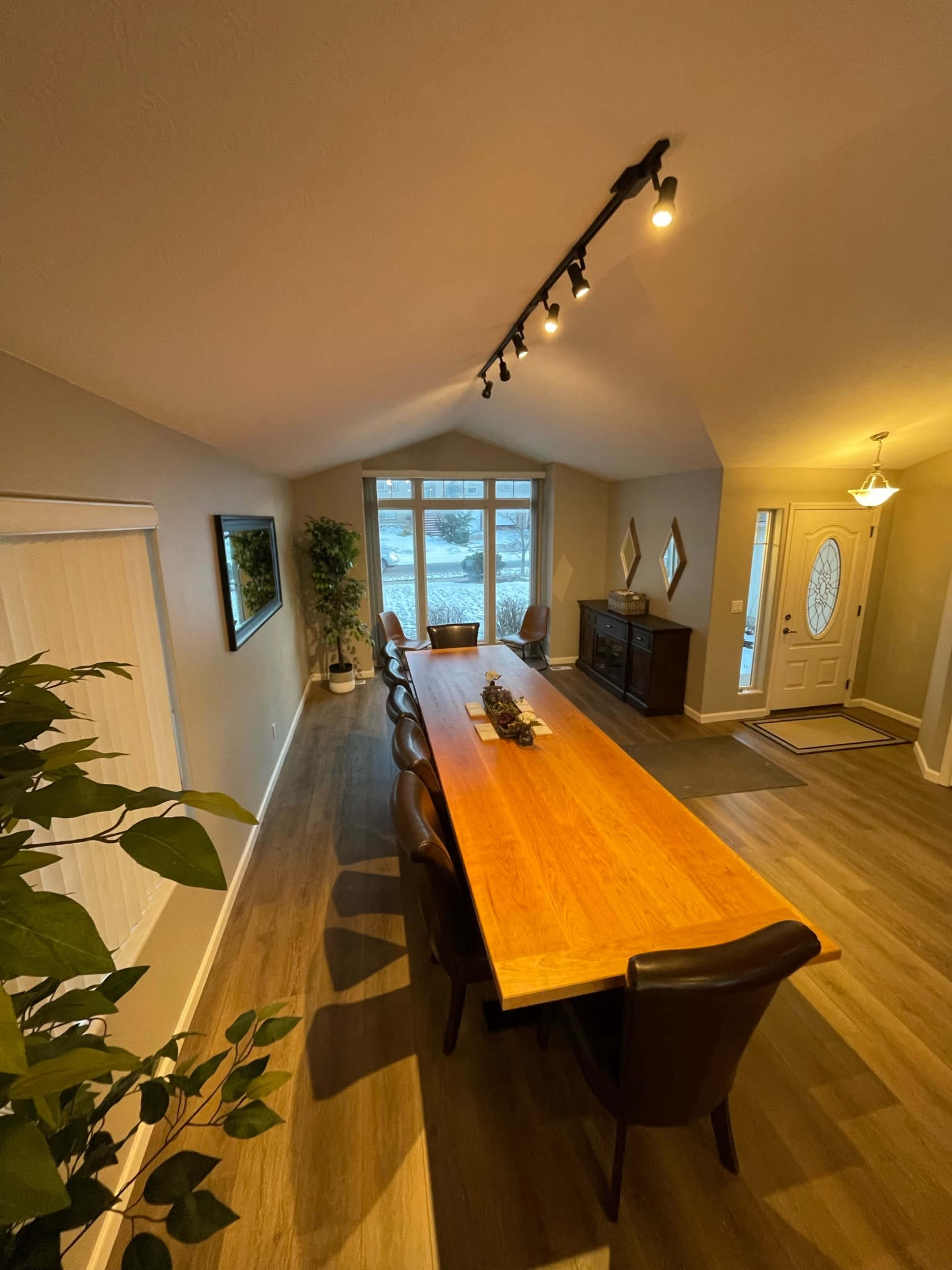 A spacious dining area features a long wooden table surrounded by leather chairs, with large windows allowing natural light to enter.